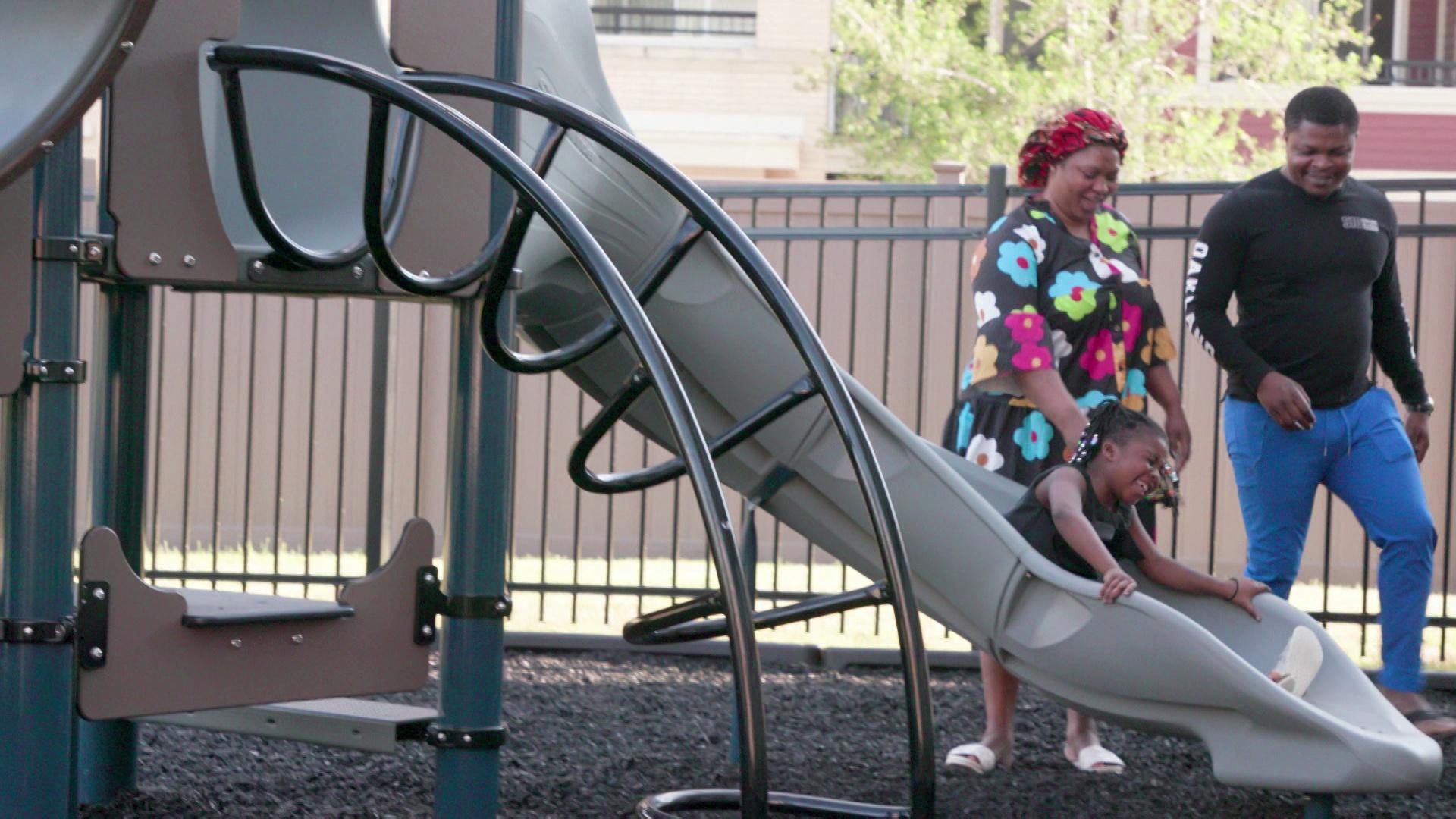 Ngwa and Stella stand on a playground while Ann Augustine sits on a slide outside with metal pipes, fencing, frees and buildings in the background.