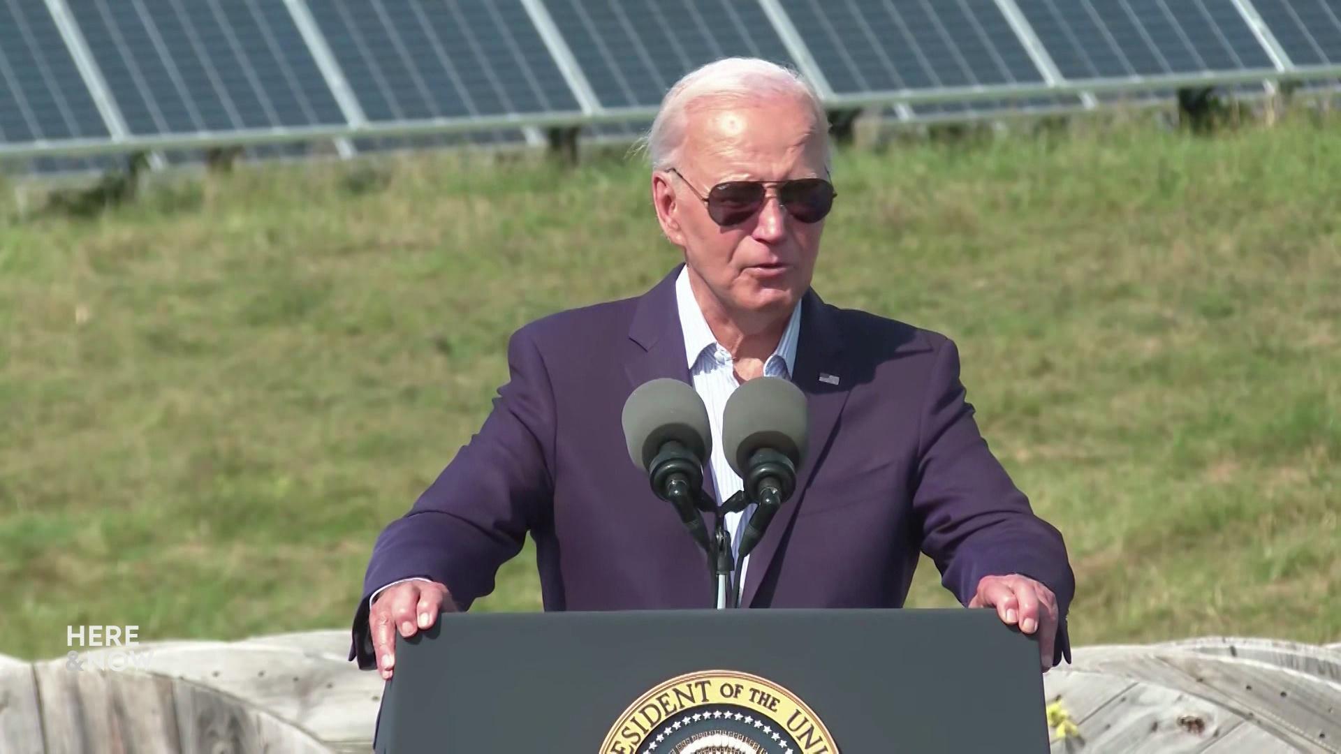 Joe Biden speaks and stands in front of a podium with two microphones with grass and solar panels in the background.