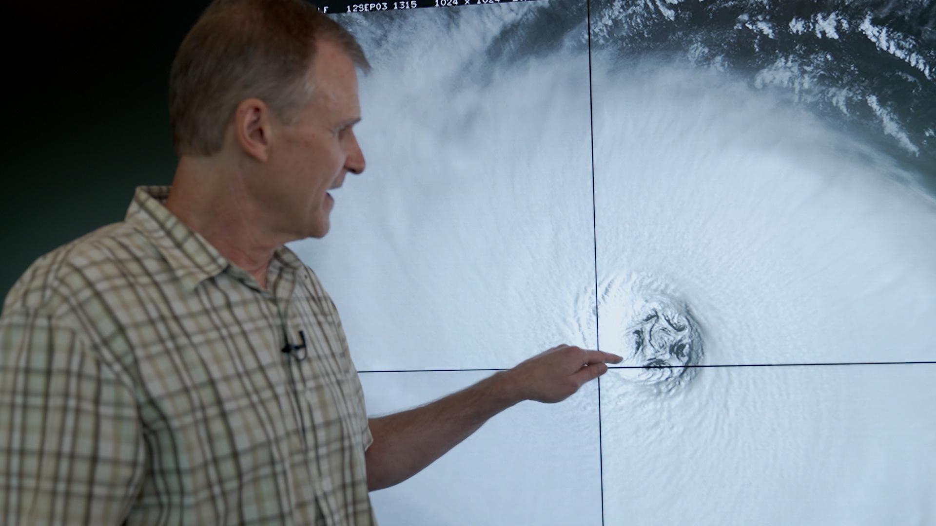 A white man pointing at the eye of a hurricane on a screen.