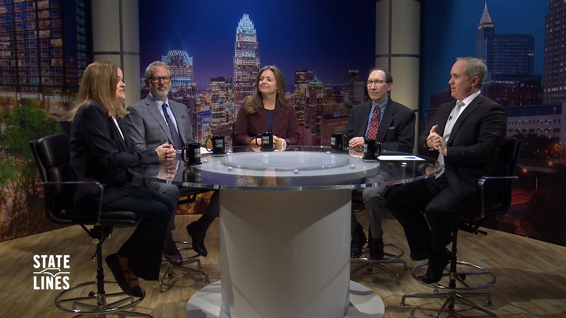 Five people — three men and two women — seated around a round studio desk during a panel discussion on the PBS NC show State Lines, with a nighttime city skyline backdrop visible behind them.