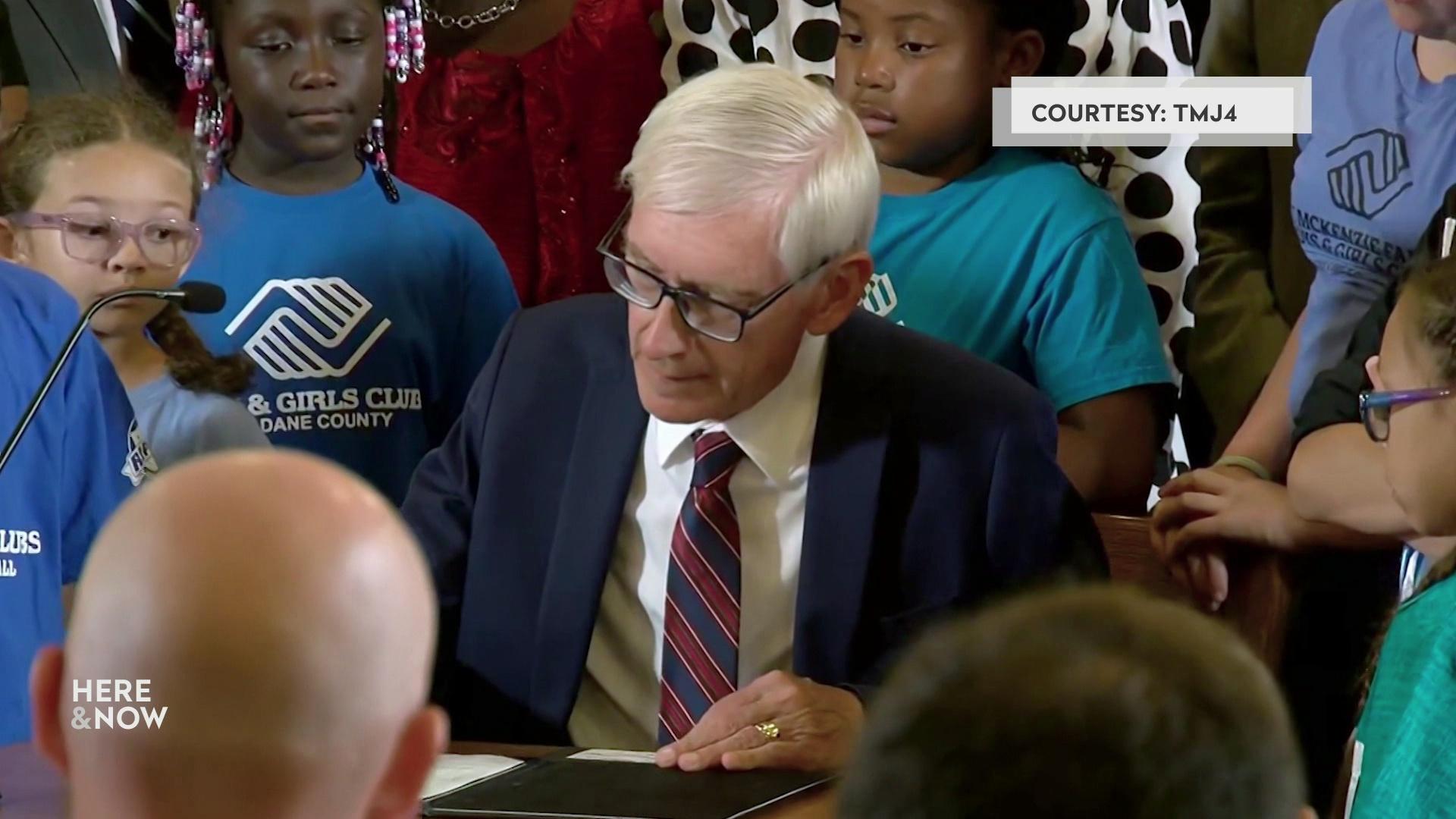 A still image shows Tony Evers signing a paper surrounded by people of various ages.