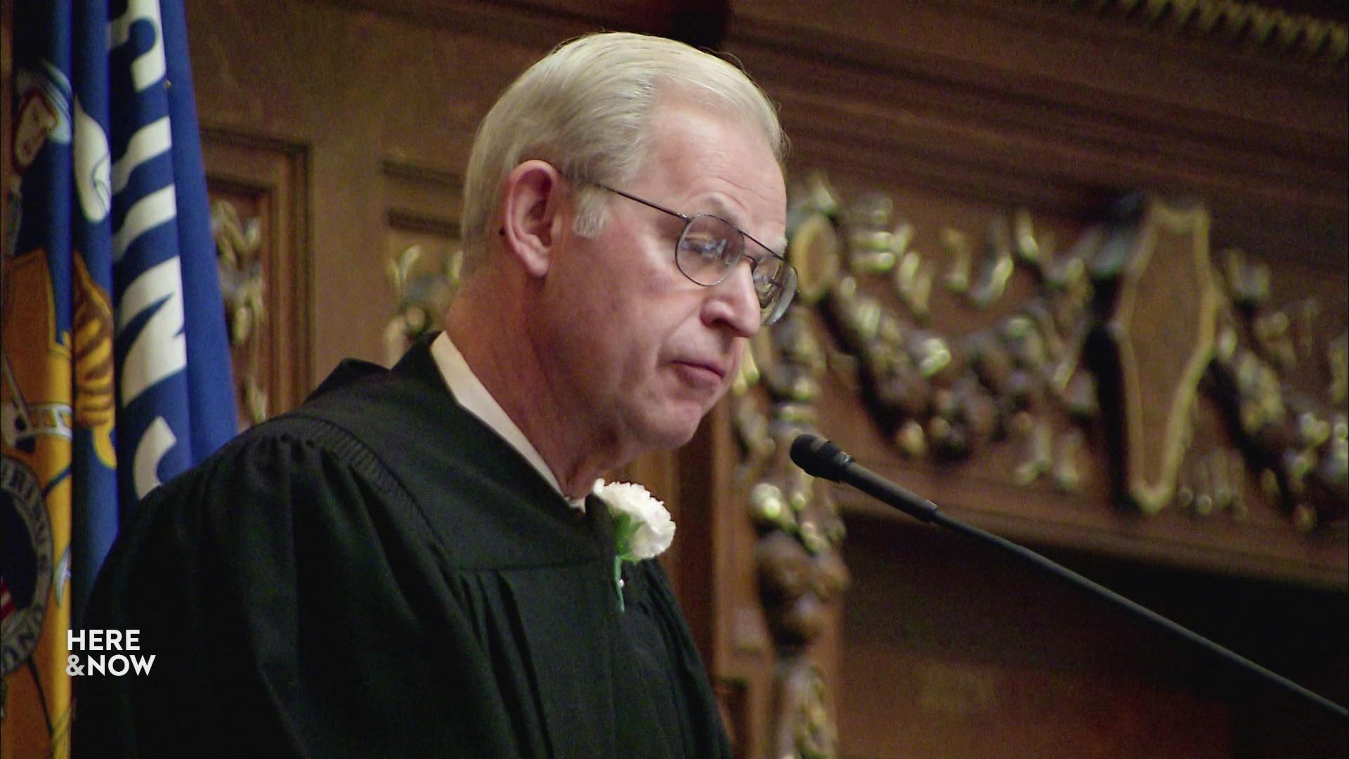 A still image shows David Prosser speaking into a microphone with wood paneling and a Wisconsin state flag in the background.