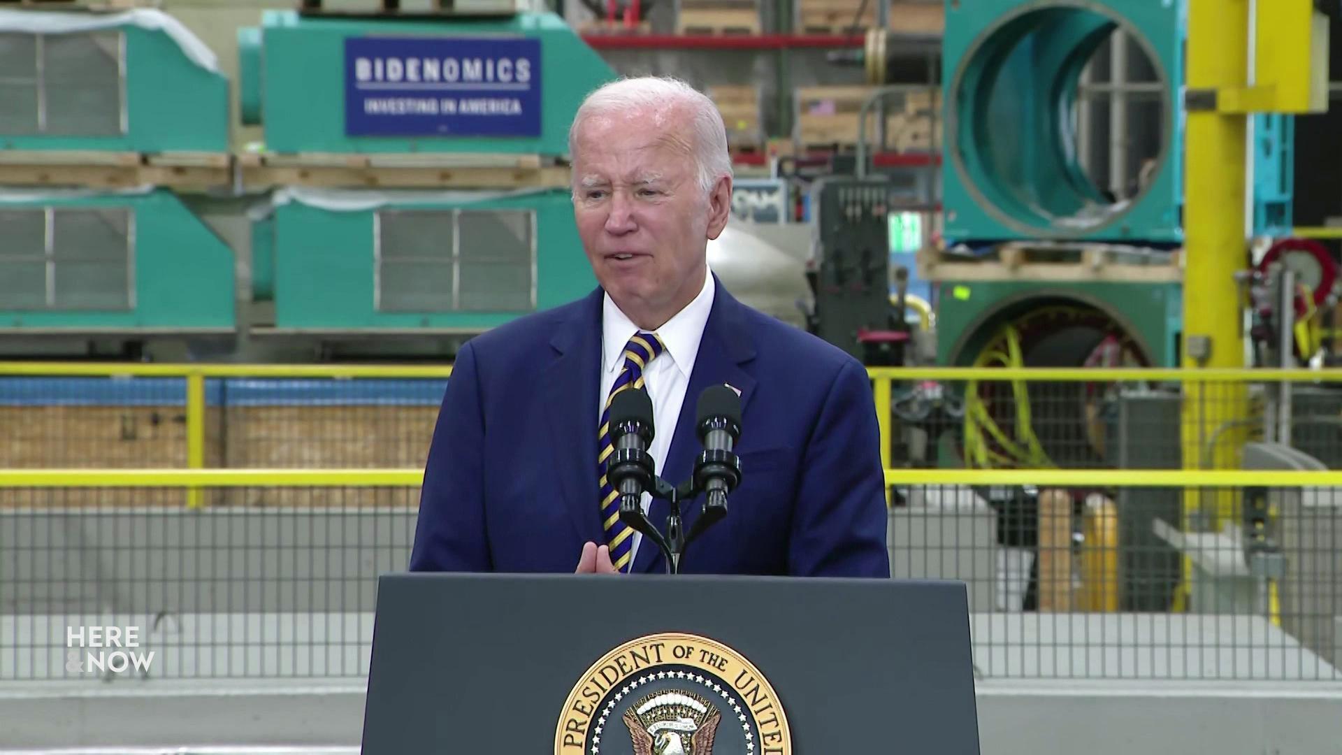 President Joe Biden stands behind a podium and microphone in an industrial warehouse with yellow and green metal and a blue poster reading 'Bidenomics.'