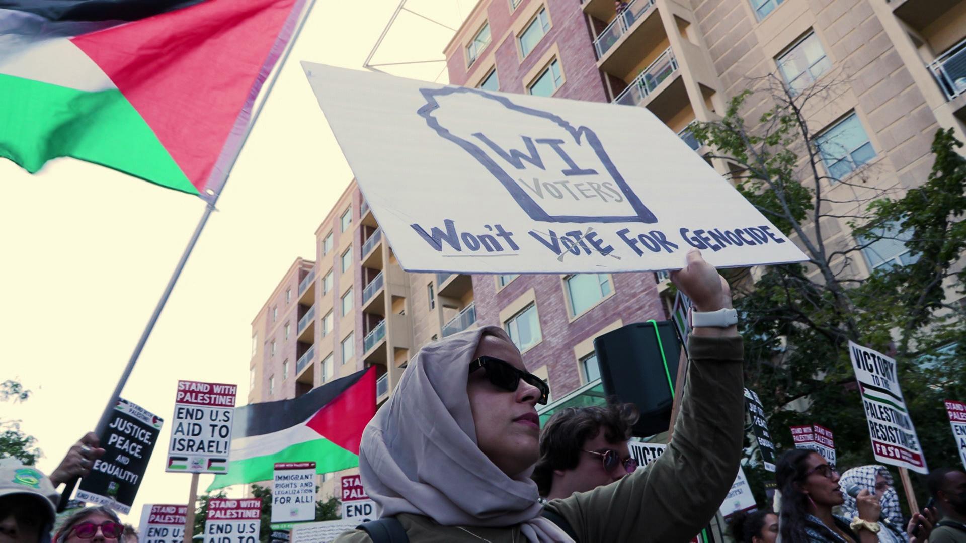 Someone holds a sign with an outline of Wisconsin that reads 'WI Voters Won't Vote For Genocide' with people walking and holding signs and Palestinian flags and trees and buildings with windows in the background.