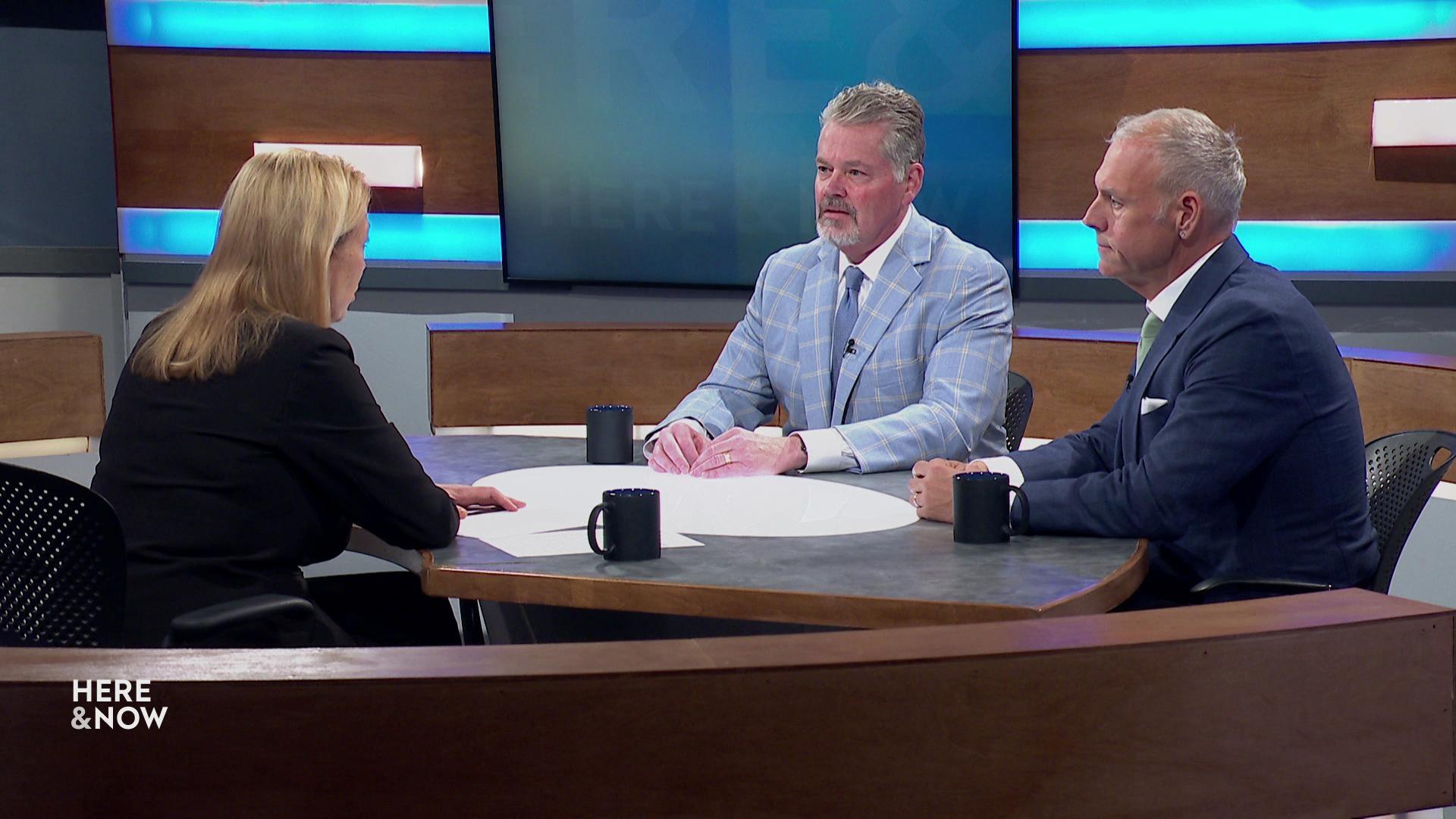A still image shows Frederica Freyberg seated across from Bill McCoshen and Scot Ross on the Here & Now set featuring wood paneling.