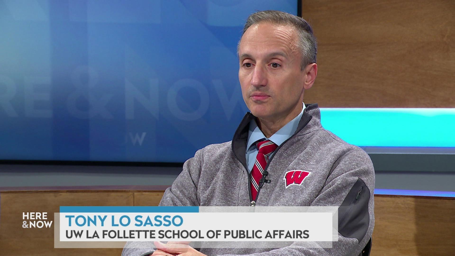 A still image shows Tony Lo Sasso seated at the 'Here & Now' set featuring wood paneling, with a graphic at bottom reading 'Tony Lo Sasso' and 'UW La Follette School of Public Affairs.'