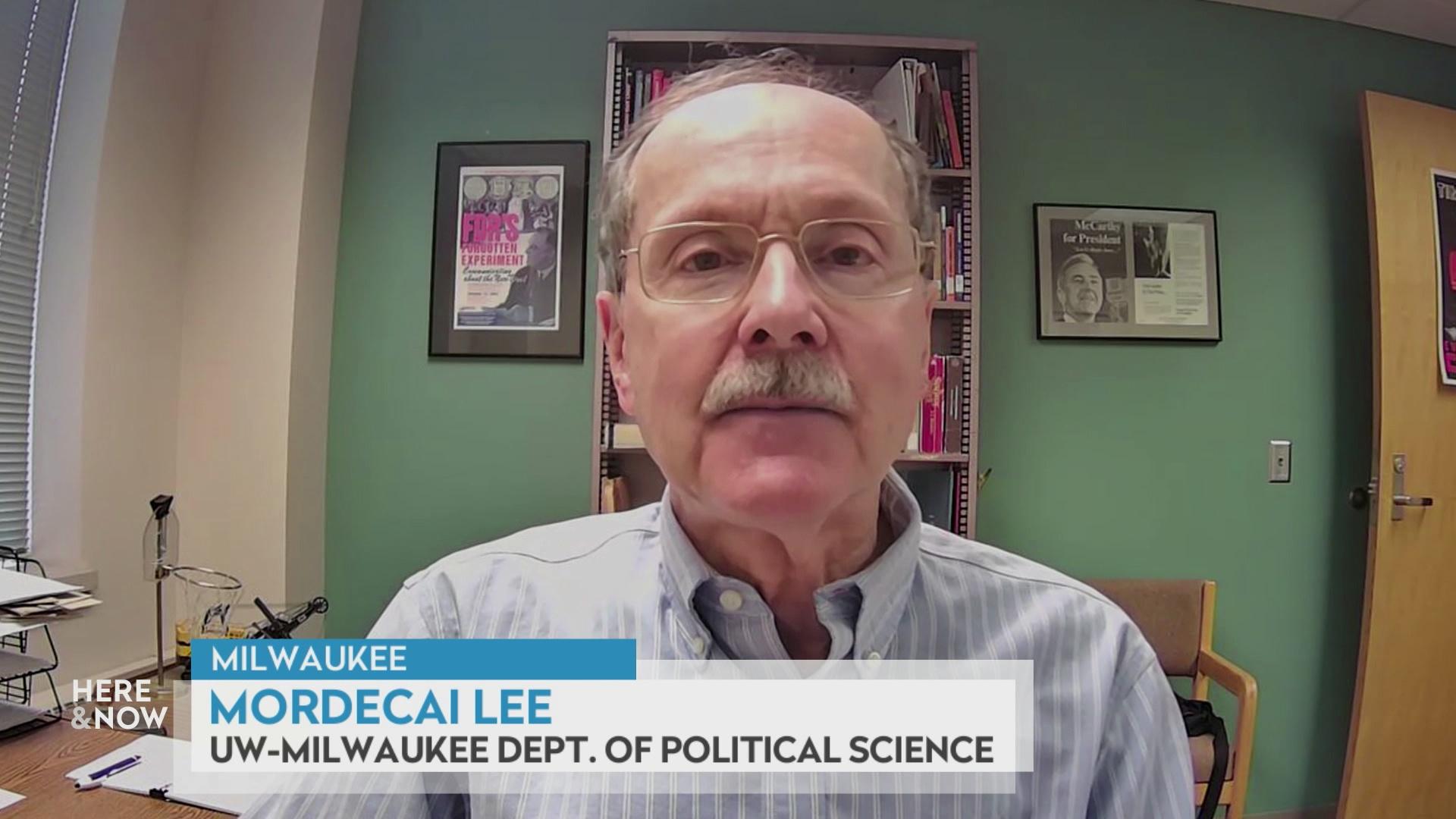 A still image from a video shows Mordecai Lee seated in front of a wall with framed art hanging on the wall on either side with a graphic at bottom reading 'Milwaukee,' 'Mordecai Lee' and 'UW-Milwaukee Dept. of Political Science.'