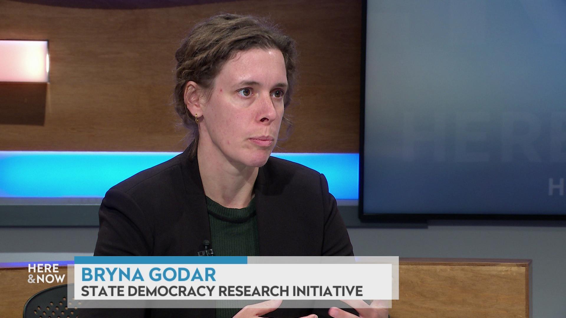 A still image shows Bryna Godar seated at the 'Here & Now' set featuring wood paneling, with a graphic at bottom reading 'Bryna Godar' and 'State Democracy Research Initiative.'