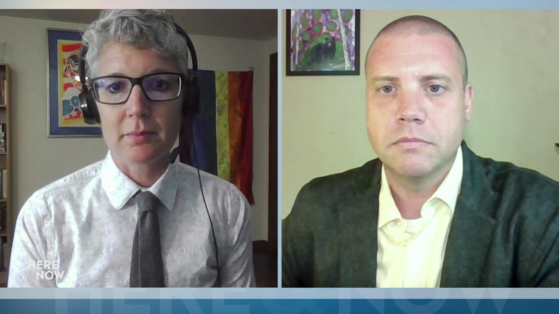 A still image from a video shows Abigail Swetz seated in front of a pride flag hung on the wall alongside Tim Peerenboom seated in front of abstract art of a bear hanging on a wall.
