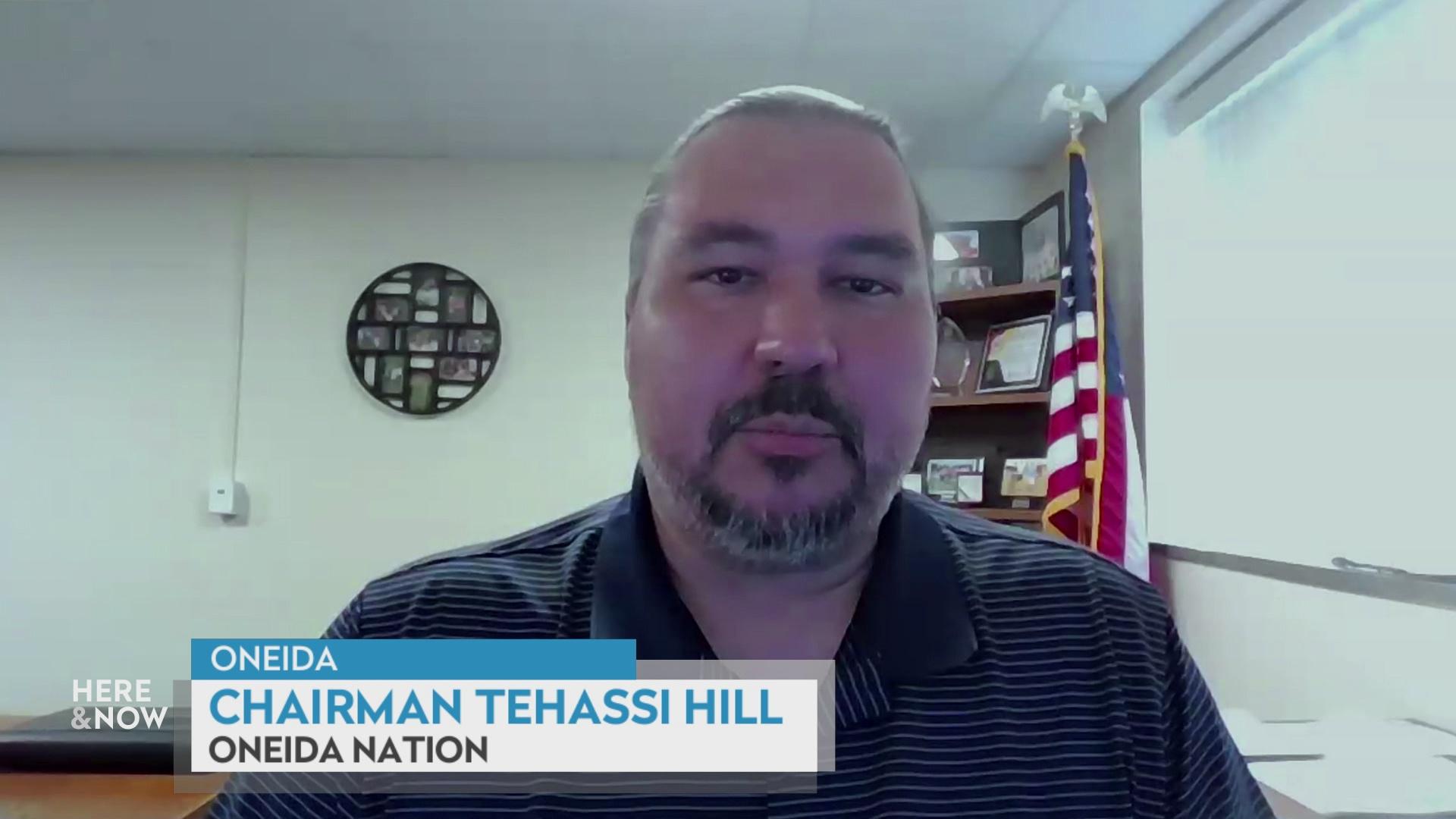 A still image from a video shows Tehassi Hill seated in front of a U.S. flag to his right and a black picture frame on a wall to his left with a graphic at bottom reading 'Oneida,' 'Chairman Tehassi Hill' and 'Oneida Nation.'