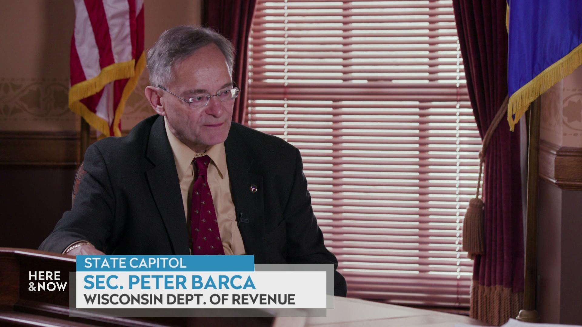 A still image from a video shows Peter Barca seated in front of a window with a U.S. flag to the left and a Wisconsin state flag to the right with a graphic at bottom reading 'Sec. Peter Barca' and 'Wisconsin Department of Revenue.'