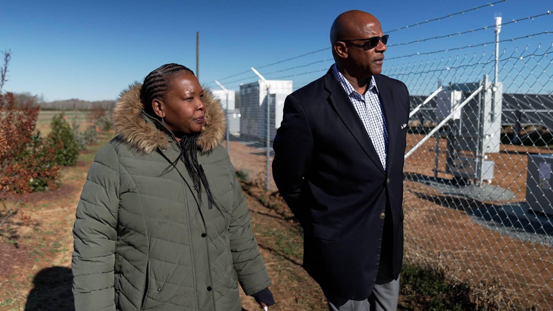 Ajulo Othlow and Marshall Cherry walking on a solar battery farm that's behind a chain-link fence.
