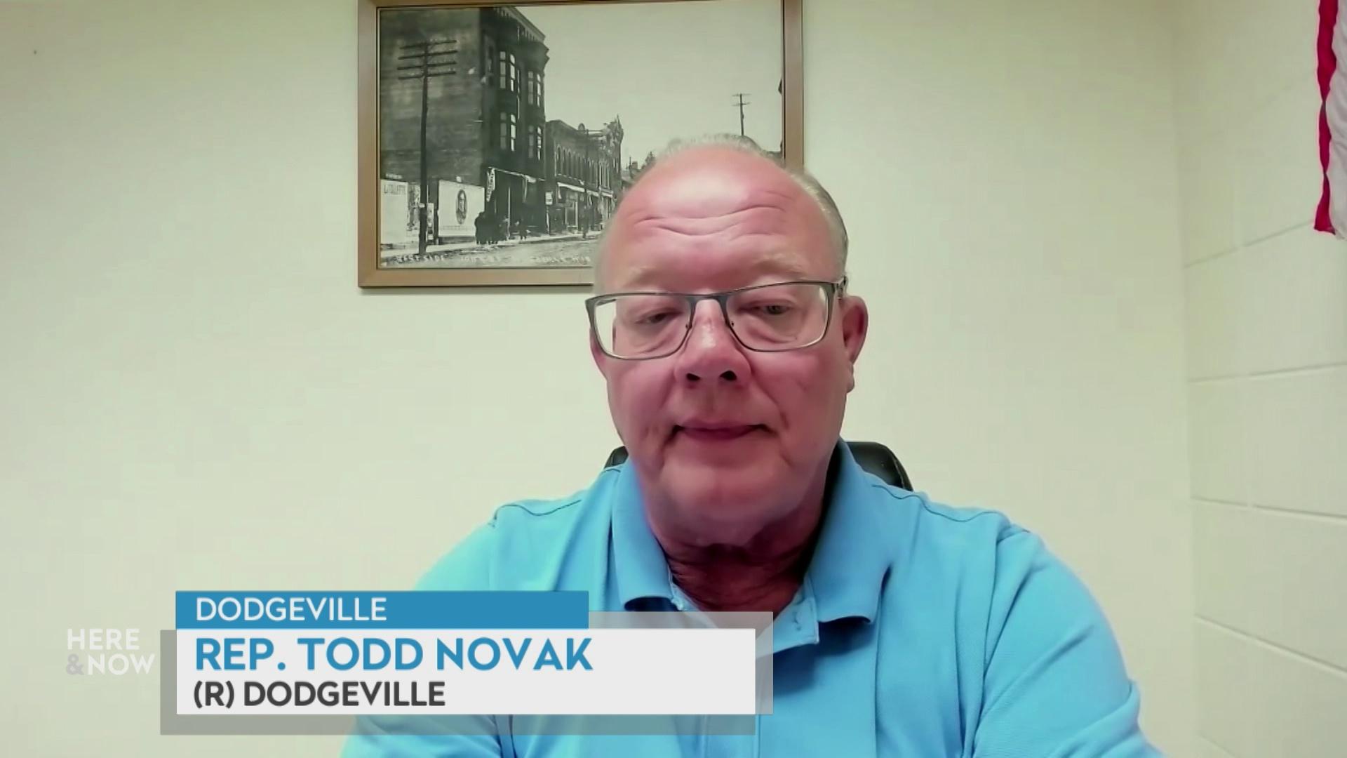 A still image from a video shows Todd Novak seated in front of a white wall with a framed black and white photograph with a graphic at bottom reading 'Dodgeville,' 'Todd Novak' and '(R) Dodgeville.'