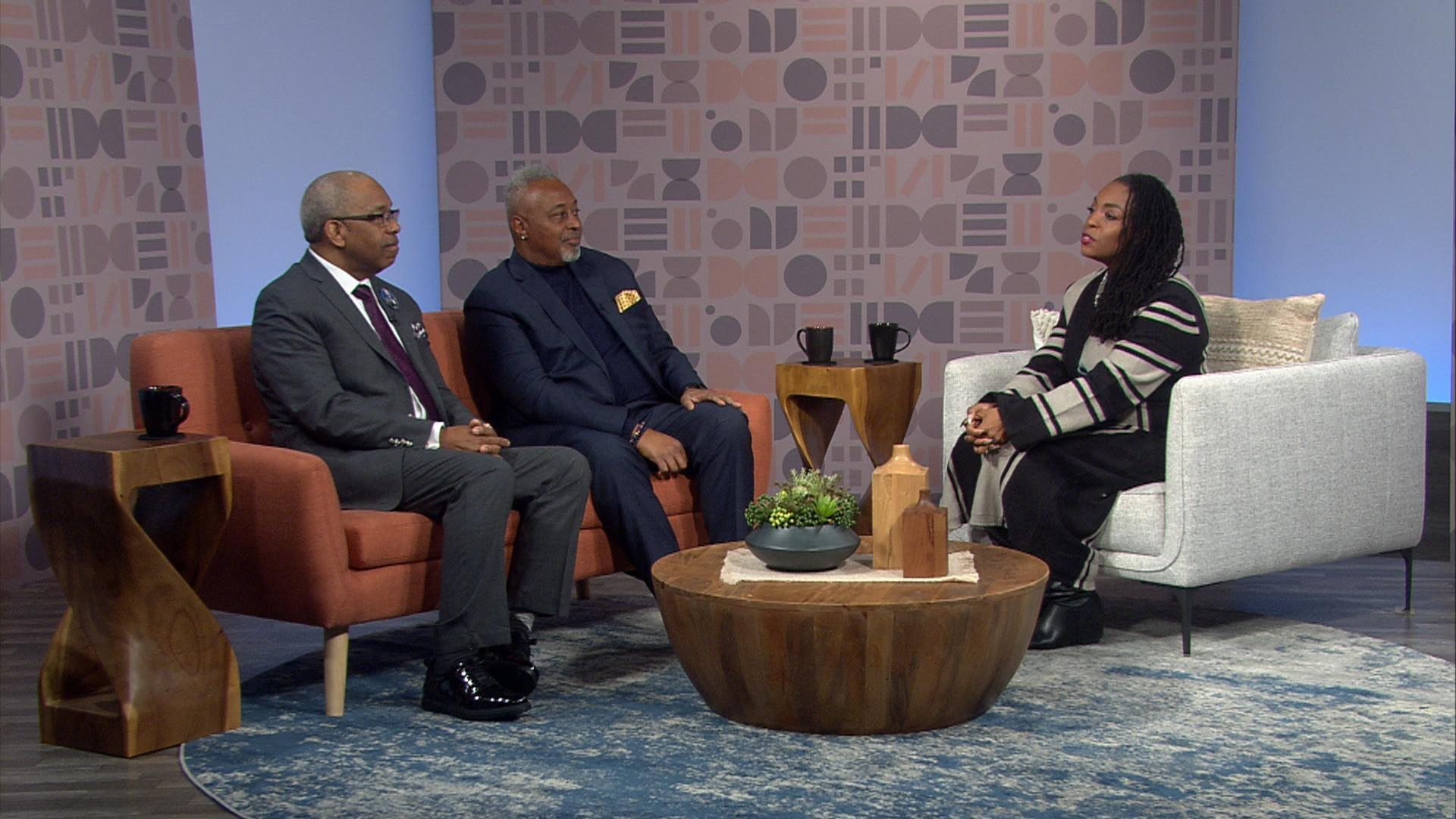 Two men and a woman seated on studio couches engaged in a conversation on a talk show set with patterned background and a wooden coffee table between them.