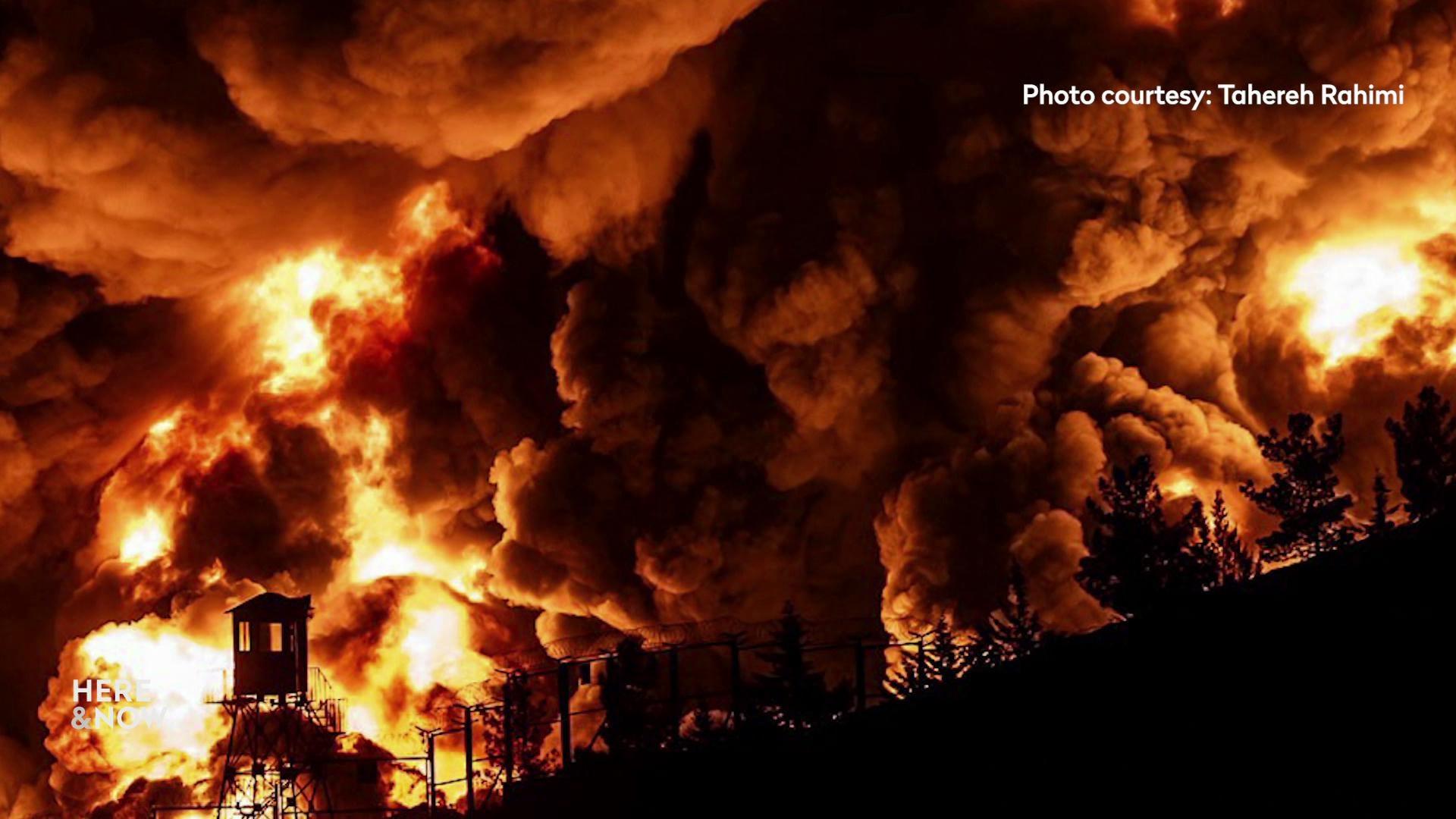 A still image shows a photo taken by Tahereh Rahimi of the aftermath of a bomb dropped on the outskirts of Tehran, fire raises high into the sky above tree lines and a watch tower, black smoke fills the rest of the sky.