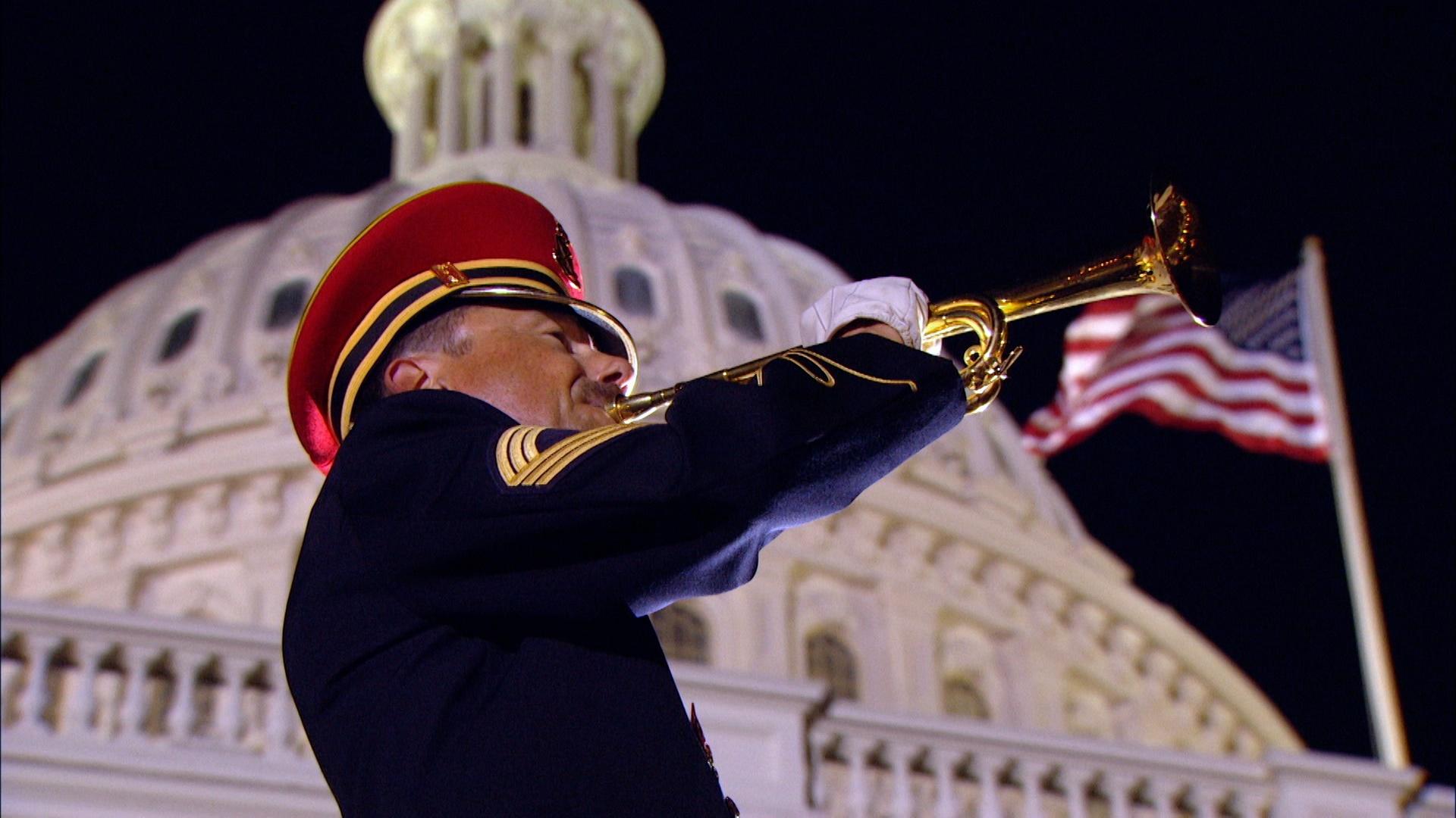 A military band member plays a trumpet with the U.S. Capitol and an American flag flying behind him.