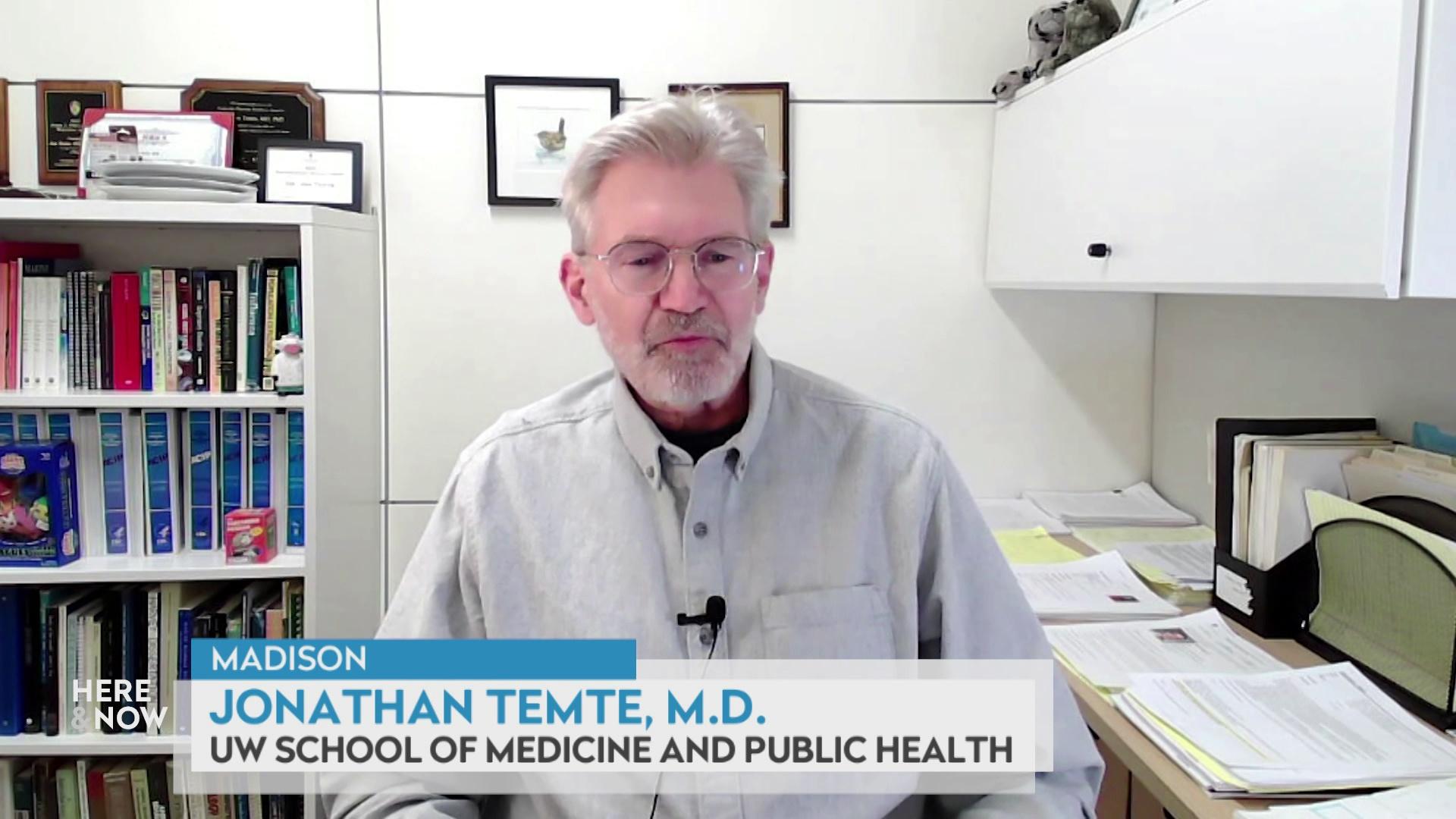 A still image from a video shows Johnathan Temte seated in front of a desk with piles of papers and a bookshelf lined with books and other items.