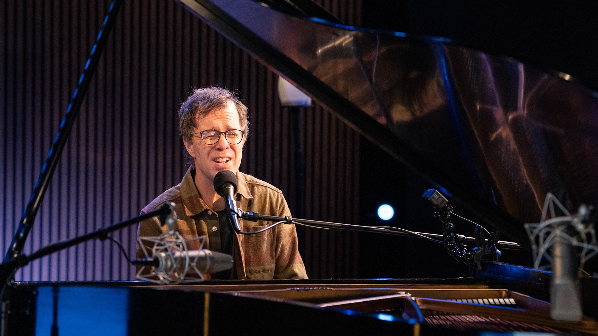Ben Folds singing into a microphone behind a grand piano.