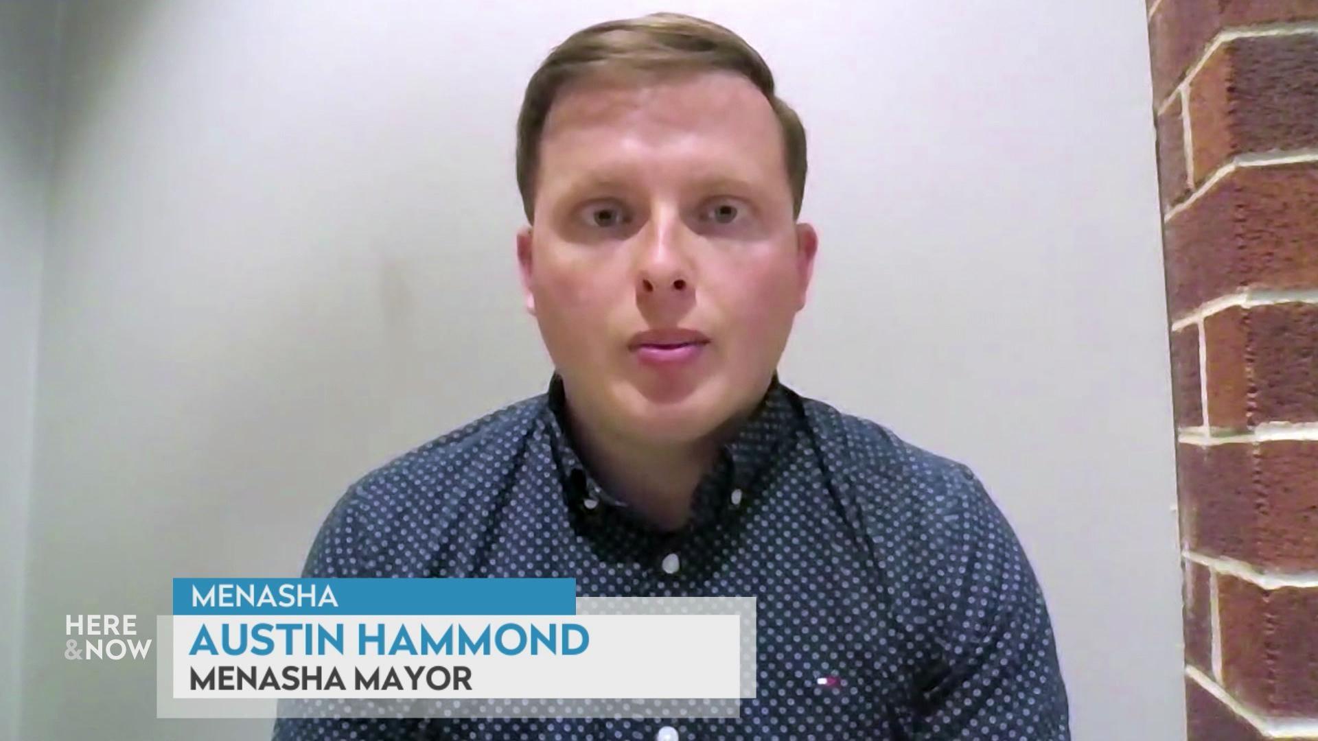 A still image shows Austin Hammond seated in front of a white wall with a brick wall to his left, with a graphic at the bottom that reads 'Menasha,' 'Austin Hammond' and 'Menasha Mayor.' A still image shows Austin Hammond seated in front of a white wall with a brick wall to his left, with a graphic at the bottom that reads 'Menasha,' 'Austin Hammond' and 'Menasha Mayor.'