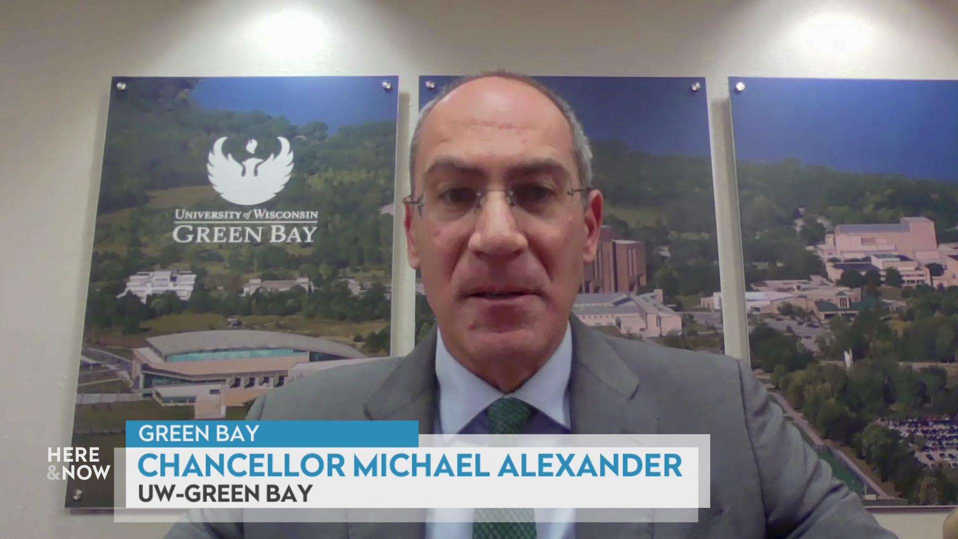 A still image from a video shows Michael Alexander seated in front of three artworks depicting trees, buildings and a stadium reading 'University of Wisconsin Green Bay' with a graphic at bottom reading 'Green Bay,' 'Chancellor Michael Alexander' and 'UW-Green Bay.'