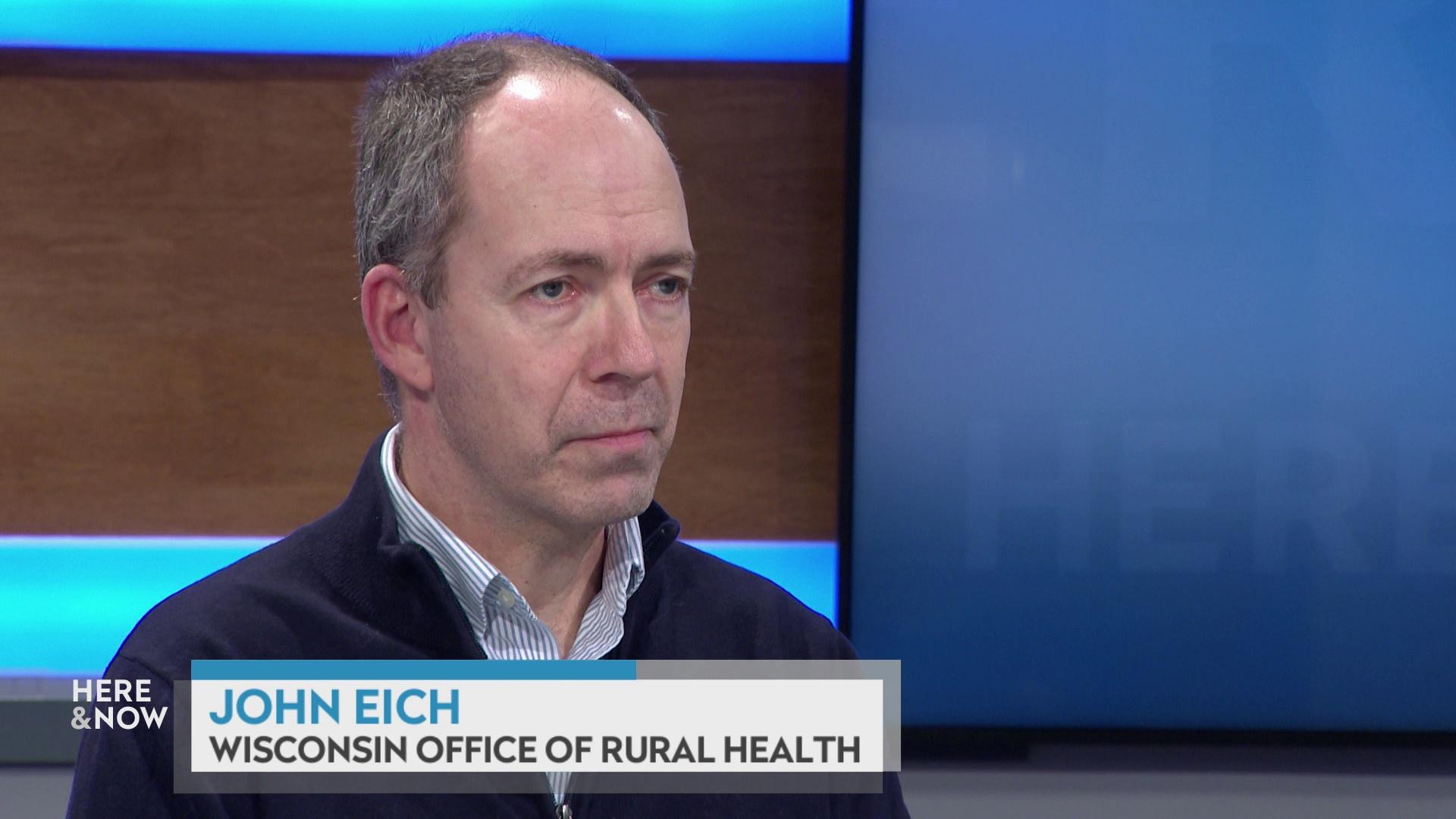 A still image shows John Eich seated at the 'Here & Now' set featuring wood paneling, with a graphic at bottom reading 'John Eich' and 'Wisconsin Office of Rural Health.'