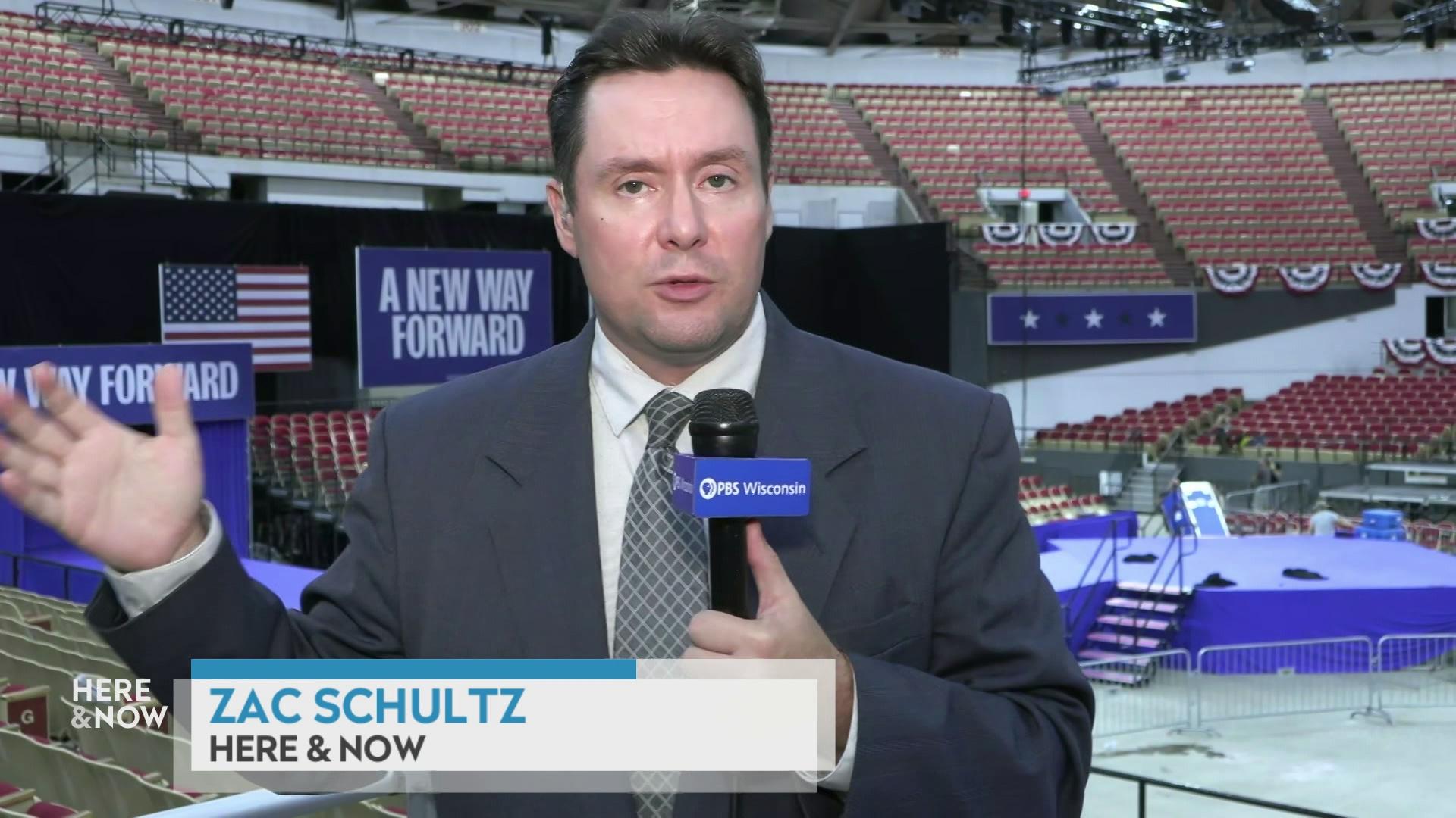 Zac Schultz speaks while standing, gesturing with his right hand and holding a microphone in the other with an empty stadium and banners reading 'A New Way Forward' in the background with a graphic at bottom reading 'Zac Schultz' and 'Here & Now.'