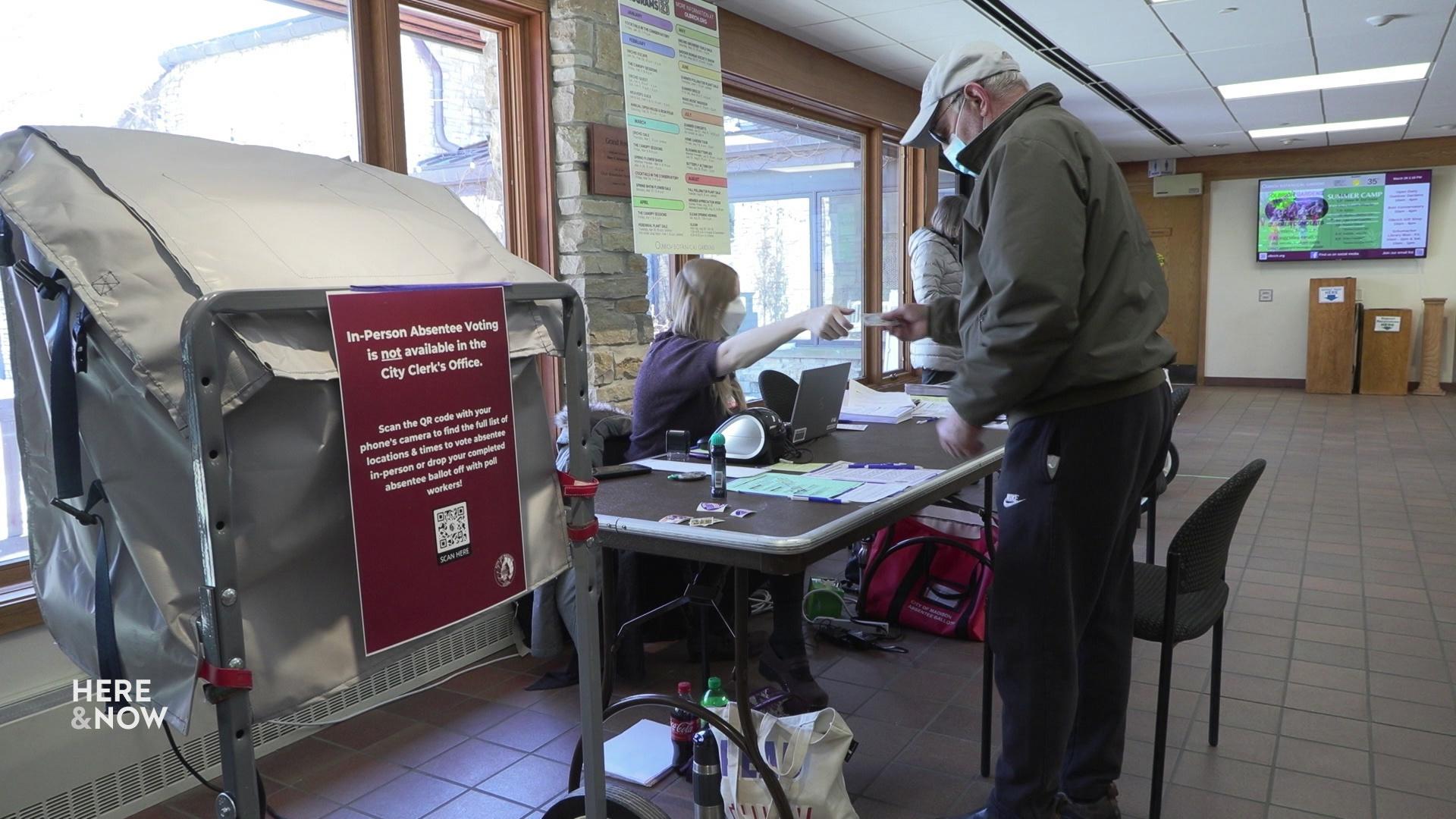A still image from a video shows a man at a polling place speaking to a clerk with a voting machine to his left.