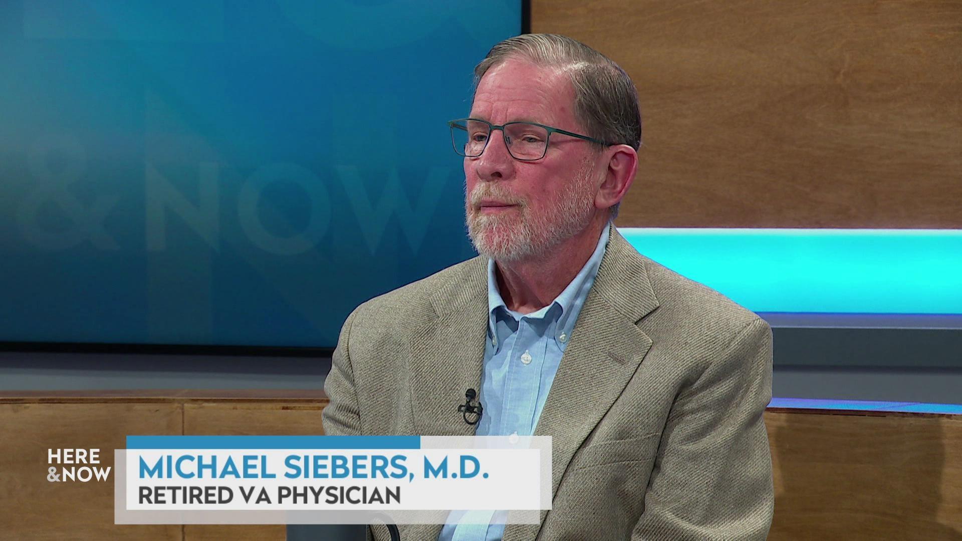 A still image shows Michael Siebers seated at the 'Here & Now' set featuring wood paneling, with a graphic at bottom reading 'Michael Siebers, M.D.' and 'Retired VA Physician'
