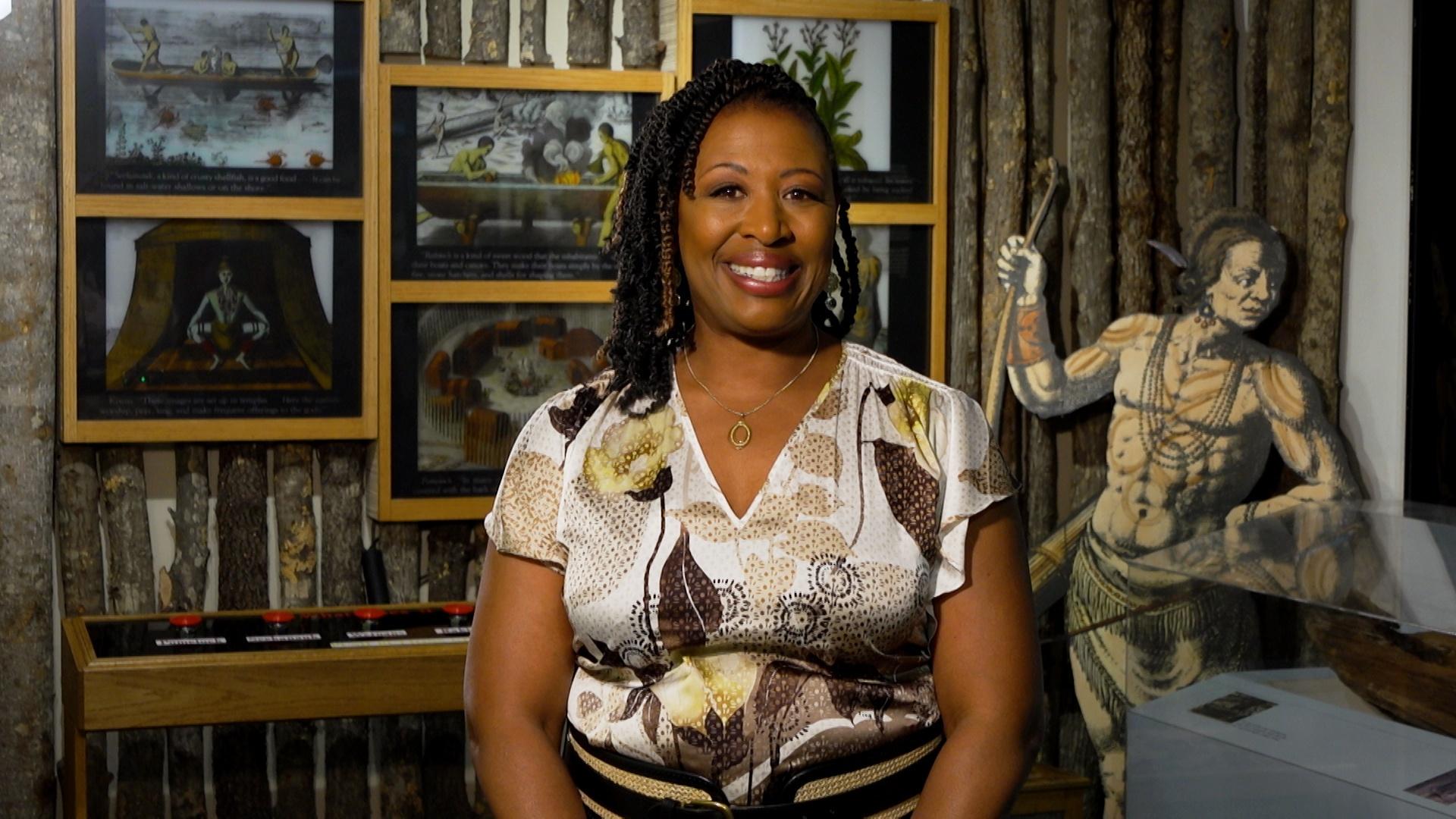 Deborah Holt Noel, host of NC Weekend in front some framed art and a cut out of a wood-print styled warrior.