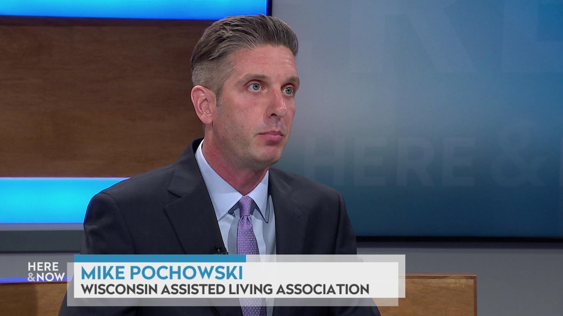 A still image shows Mike Pochowski seated at the 'Here & Now' set featuring wood paneling, with a graphic at bottom reading 'Mike Pochowski' and 'Wisconsin Assisted Living Association.'