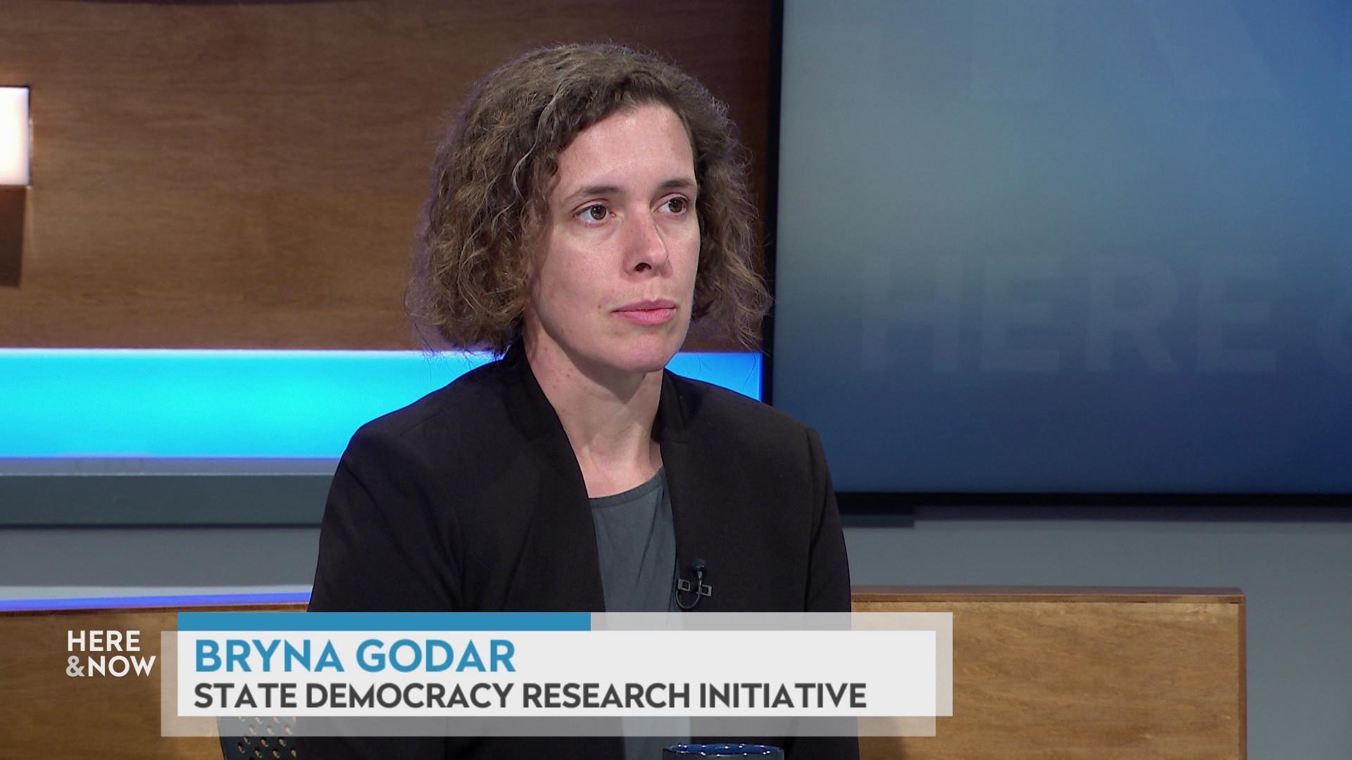 A still image shows Bryna Godar seated at the 'Here & Now' set featuring wood paneling, with a graphic at bottom reading 'Bryna Godar' and 'State Democracy Research Initiative.'