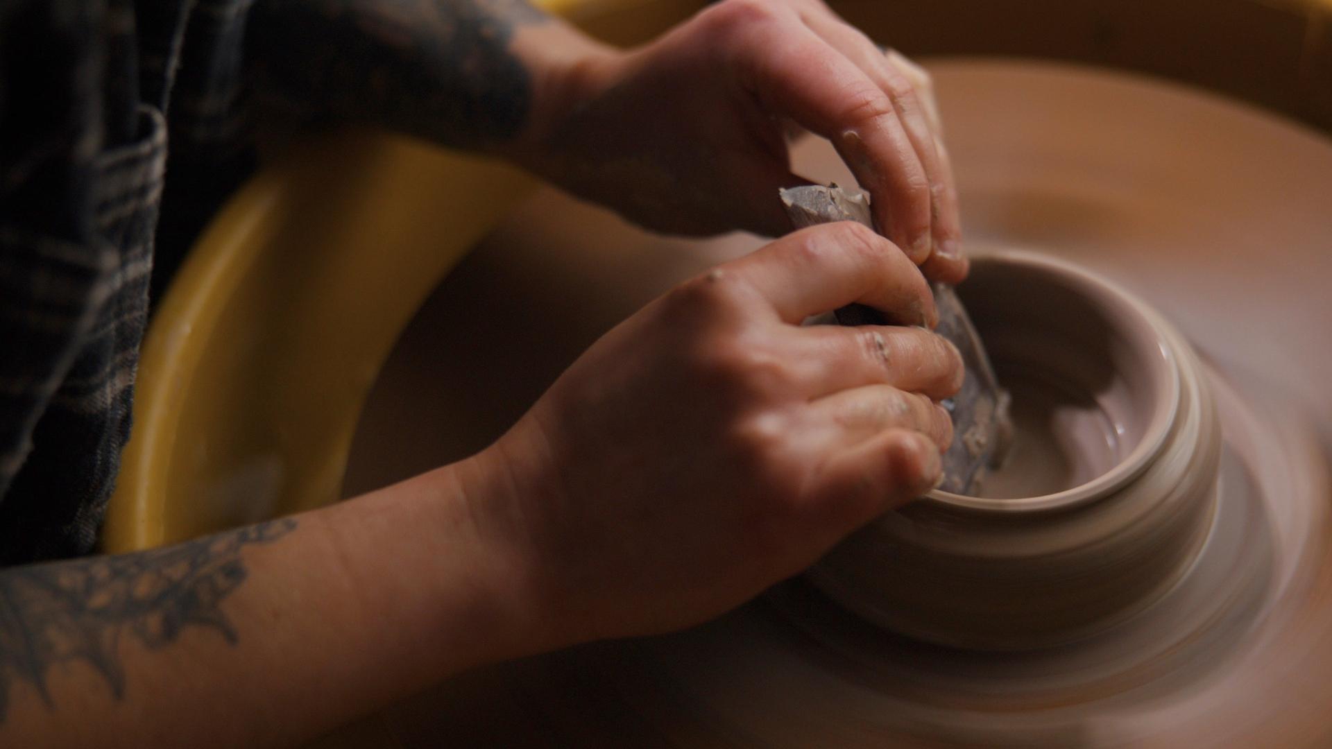 The hands of a person forming the start of a vase on a potter wheel.