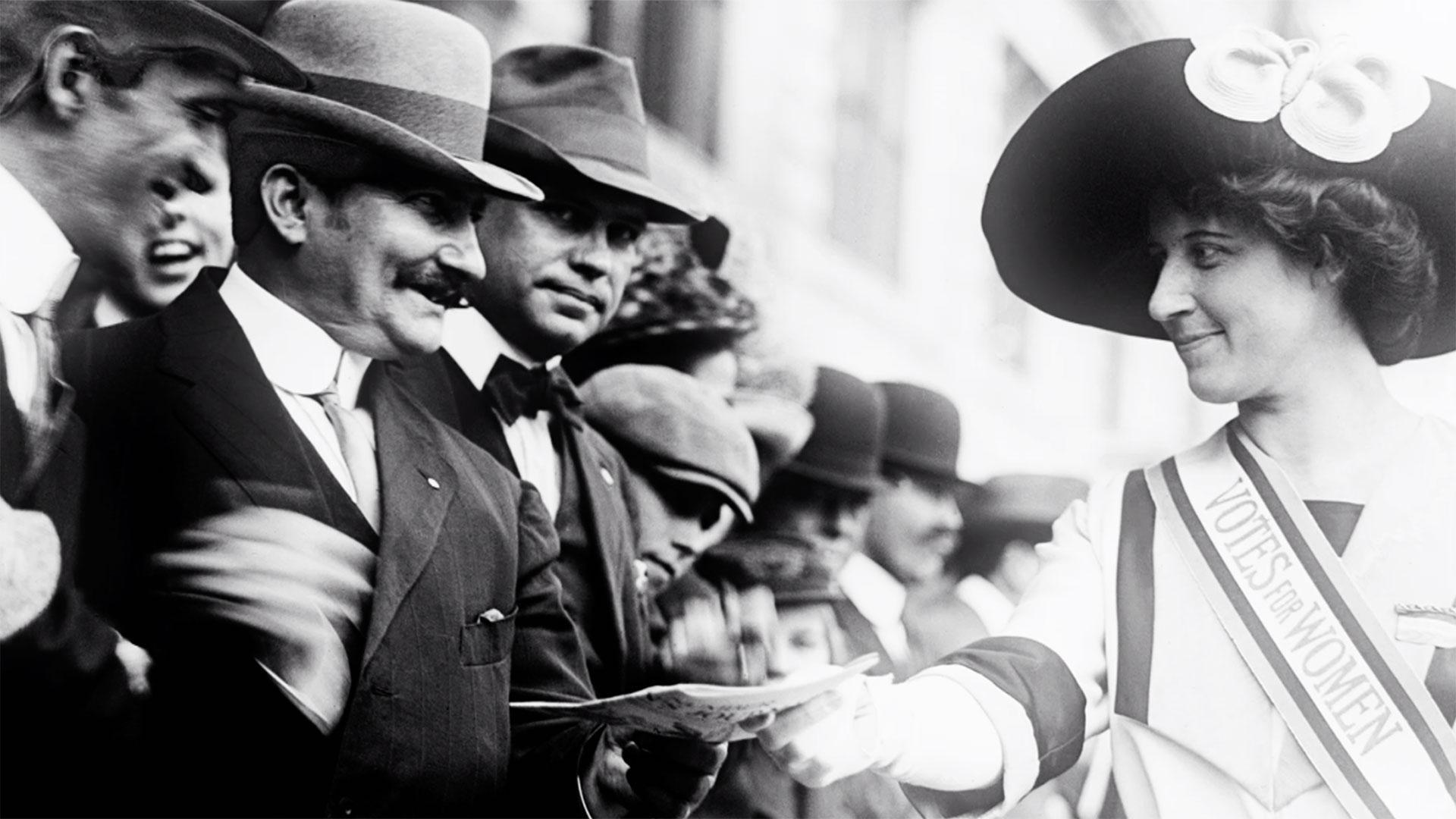 Woman with Votes for Women sash walks by a crowd