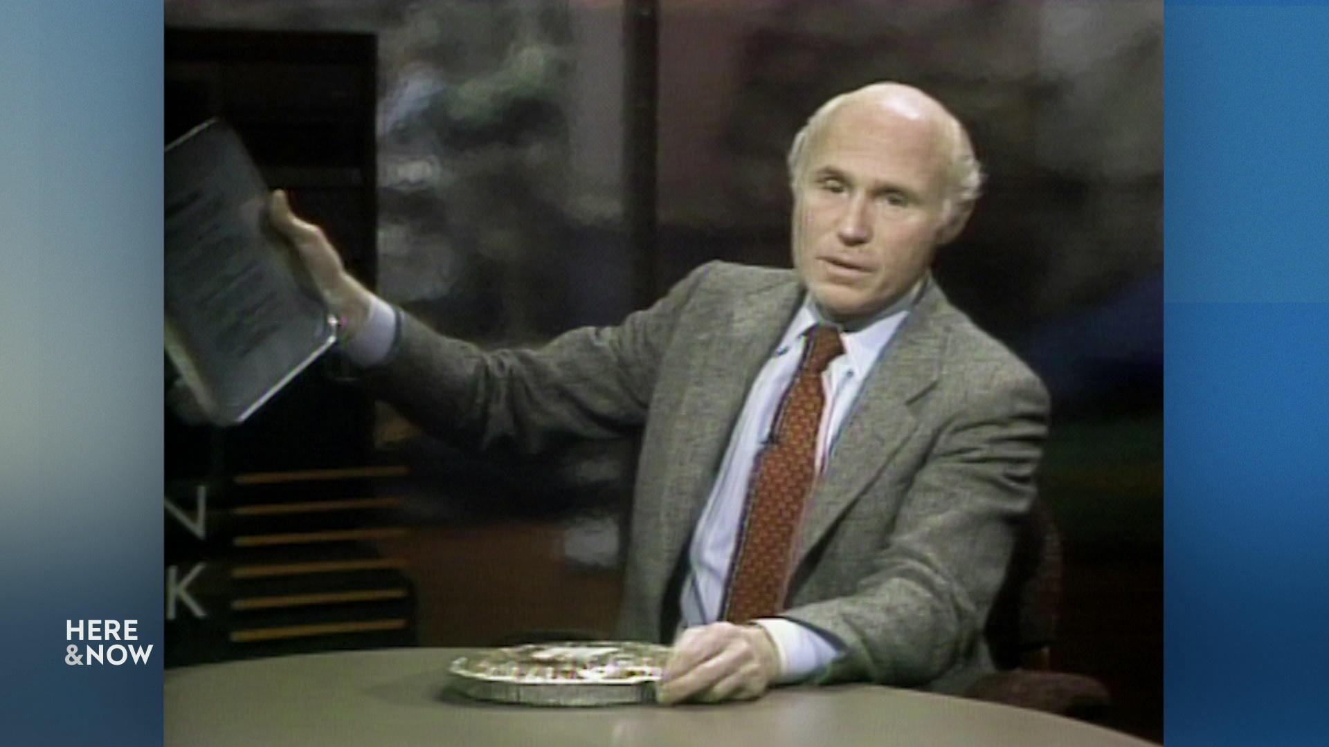 A still image from a video shows Herb Kohl holding a stack of papers behind a desk.