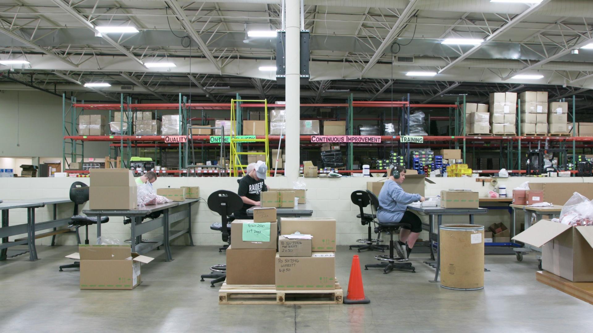 Rows of people sit in chairs at desks with piles of cardboard boxes on the side inside a large warehouse with ceiling-high shelves holding bosses and items in the background.