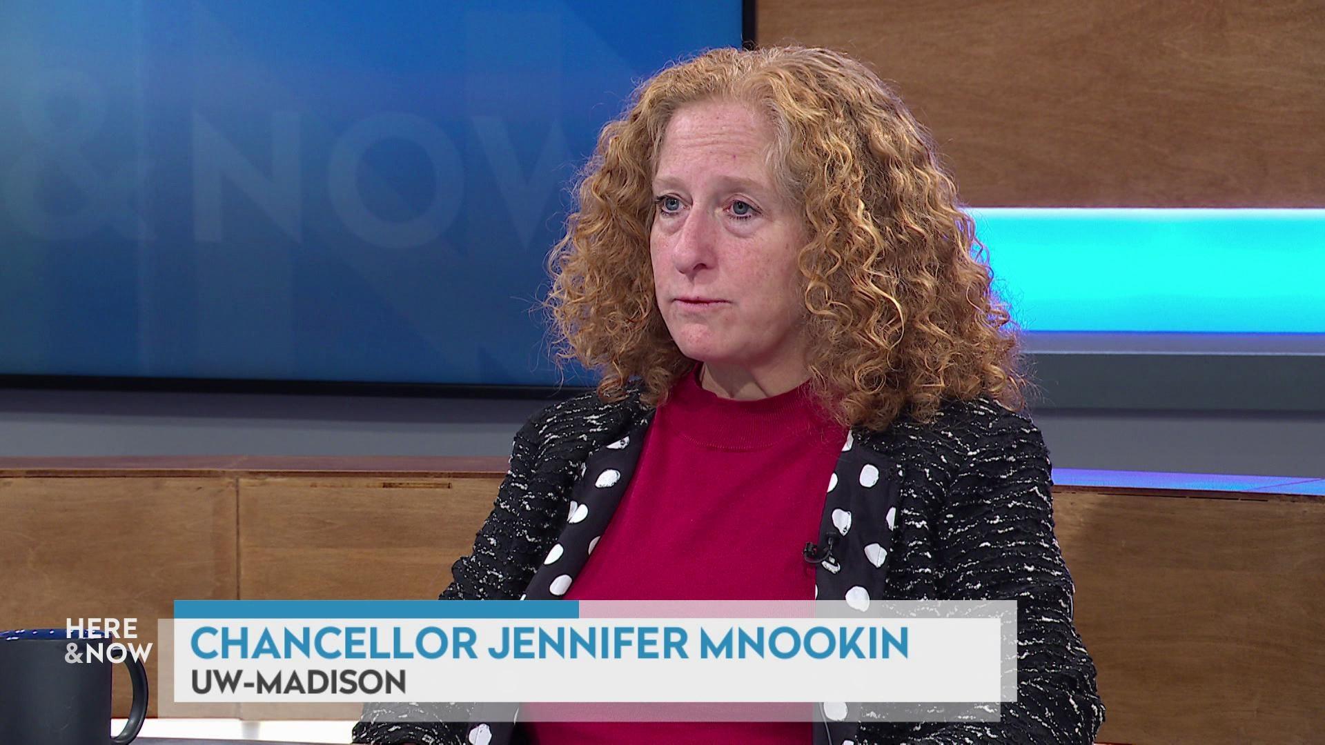 A still image shows Jennifer Mnookin seated at the 'Here & Now' set featuring wood paneling, with a graphic at bottom reading 'Chancellor Jennifer Mnookin' and 'UW-Madison.' A still image shows Jennifer Mnookin seated at the 'Here & Now' set featuring wood paneling, with a graphic at bottom reading 'Chancellor Jennifer Mnookin' and 'UW-Madison.'