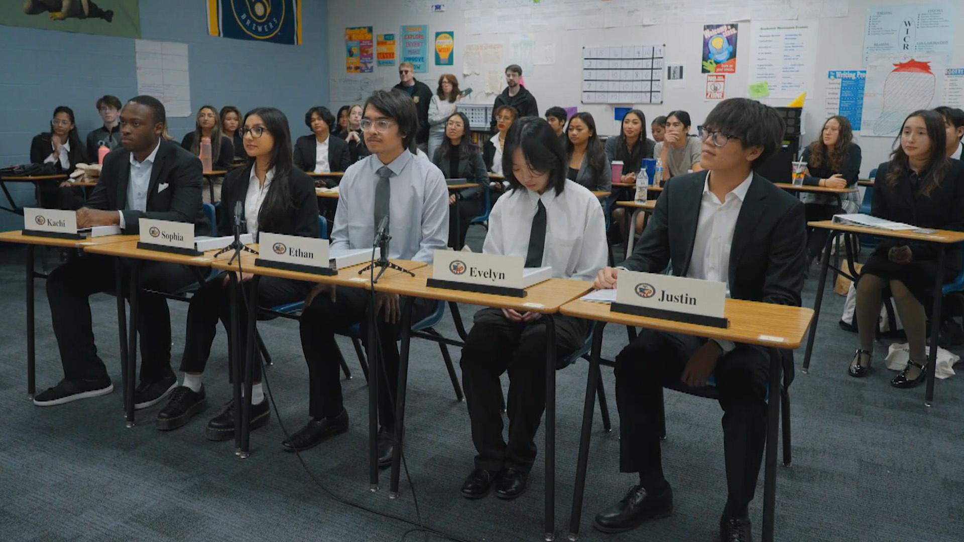 Students sitting in their desks dressed in suits with name cards in front of them.