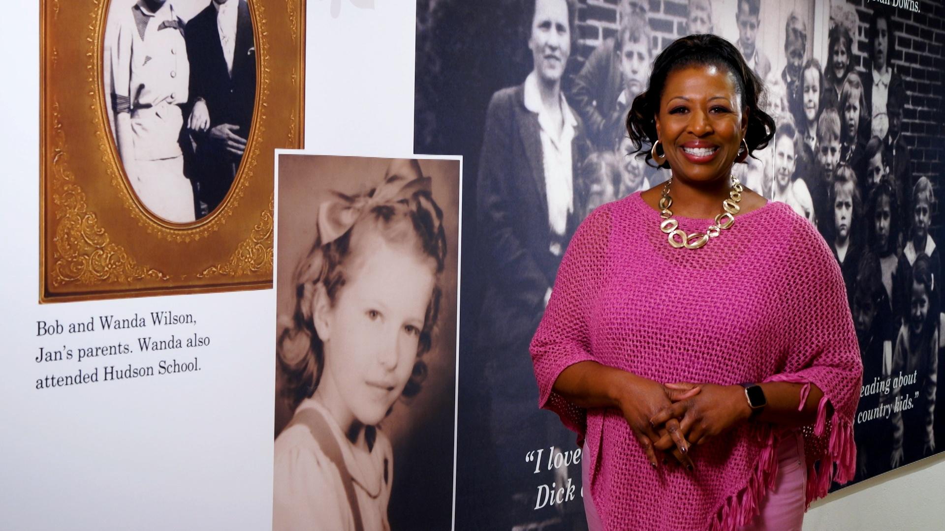 Host of NC Weekend, Deborah Holt Noel, wearing a bright pink sweater and a large circular necklace while standing in front of a wall full of historical images and text of Jan Karon in the Mitford Museum of Hudson, North Carolina.