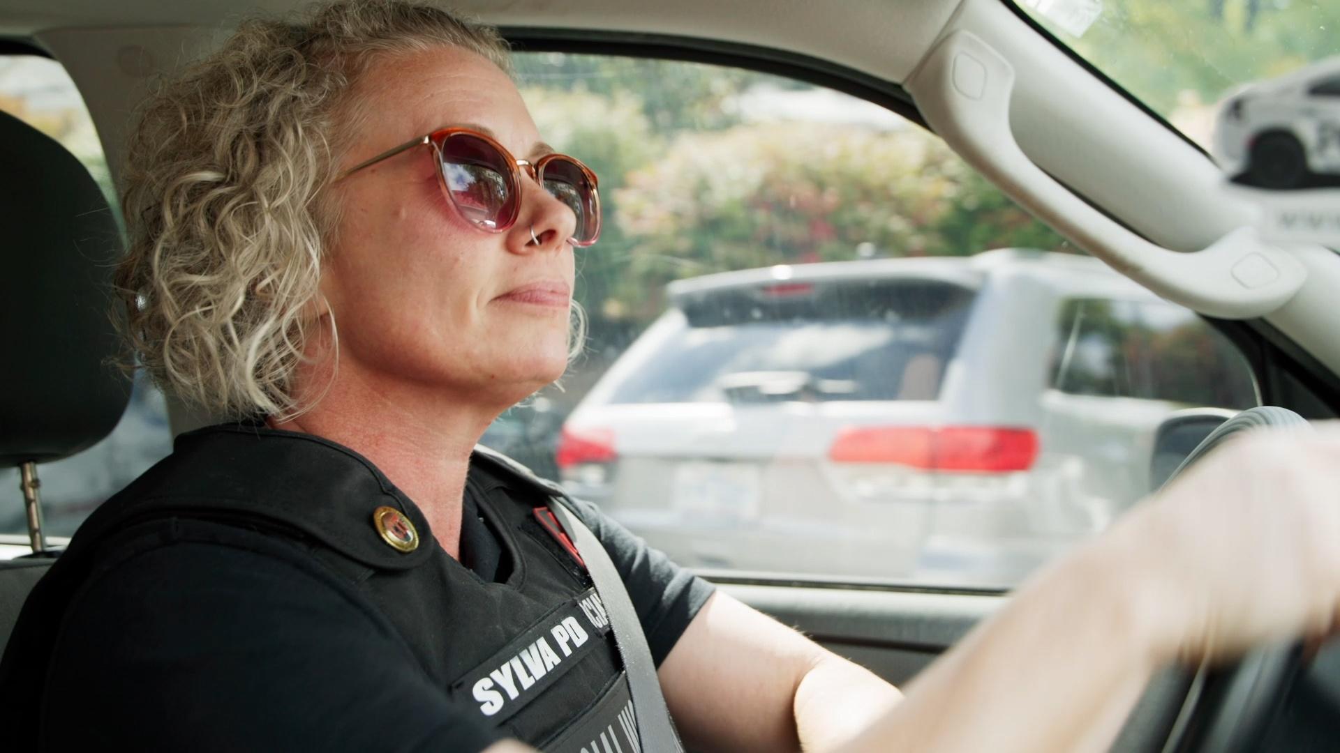 A social worker intern in a bullet proof vest and sunglasses while driving a car.