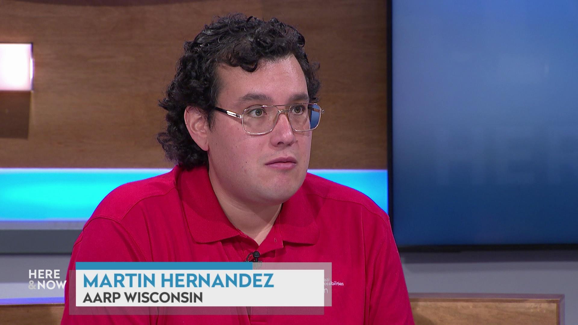 A still image shows Martin Hernandez seated at the 'Here & Now' set featuring wood paneling, with a graphic at bottom reading 'Martin Hernandez' and 'AARP Wisconsin.'