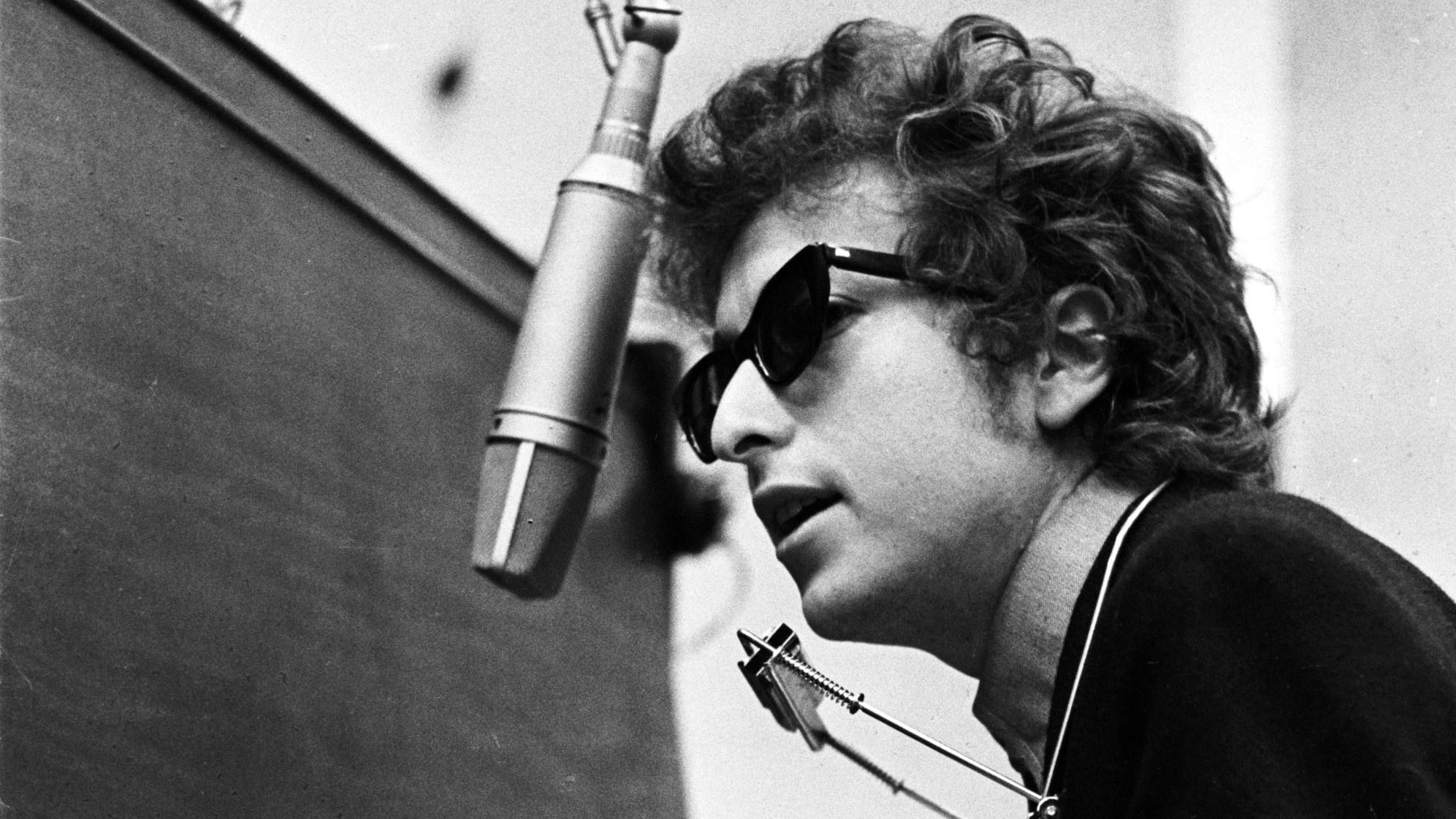 A young Bob Dylan wearing sunglasses with harmonica and microphone