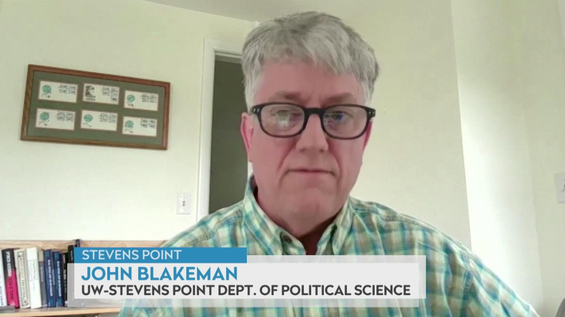 A still image from a video shows John Blakeman seated in front of a doorframe and framed artwork to his left, with a graphic at bottom reading 'Stevens Point,' 'John Blakeman' and 'UW-Stevens Point Dept. of Political Science.'