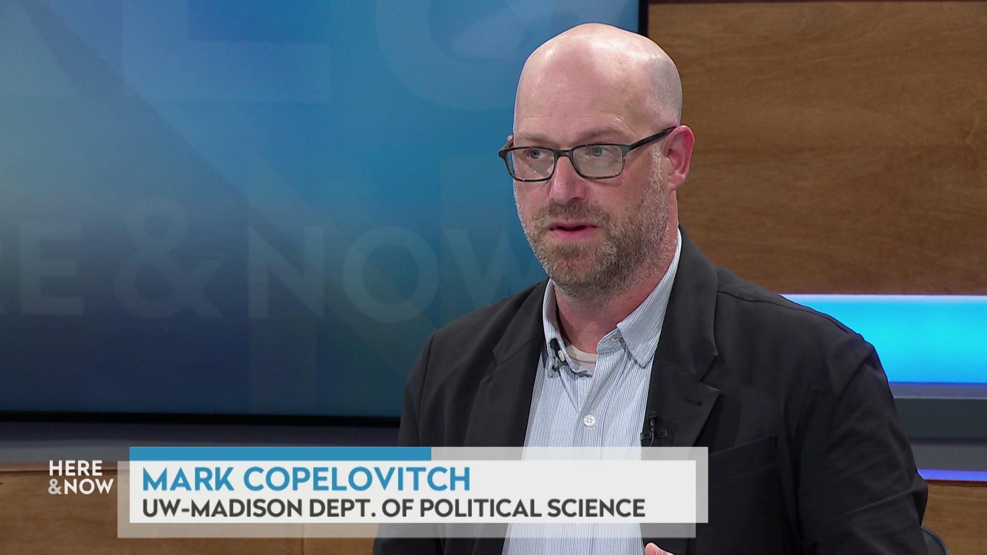 A still image shows Mark Copelovitch seated at the 'Here & Now' set featuring wood paneling, with a graphic at bottom reading 'Mark Copelovitch' and 'UW-Madison Dept. of Political Science.'