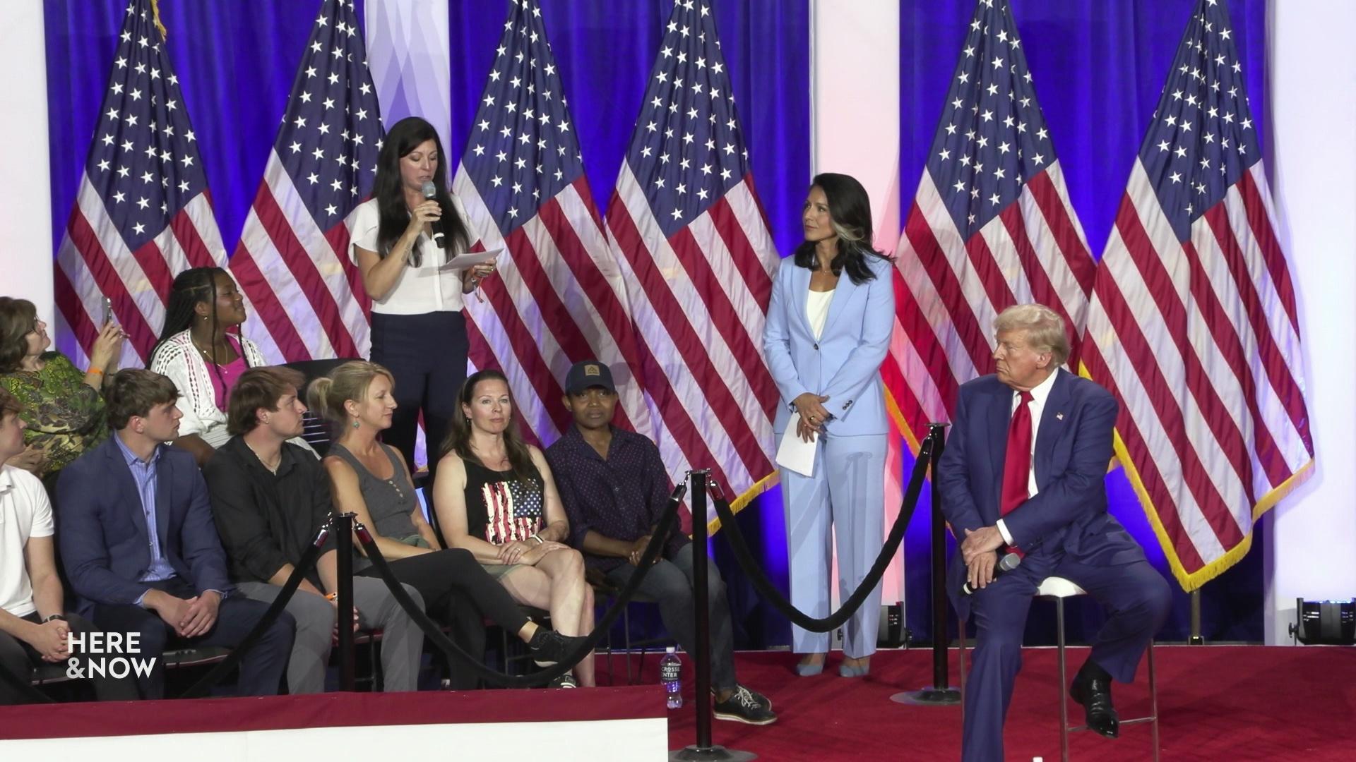 A still image shows Donald Trump seated on stage facing two rows of people seated behind a rope with one person standing and holding a microphone, another person standing and six U.S. flags in the background.