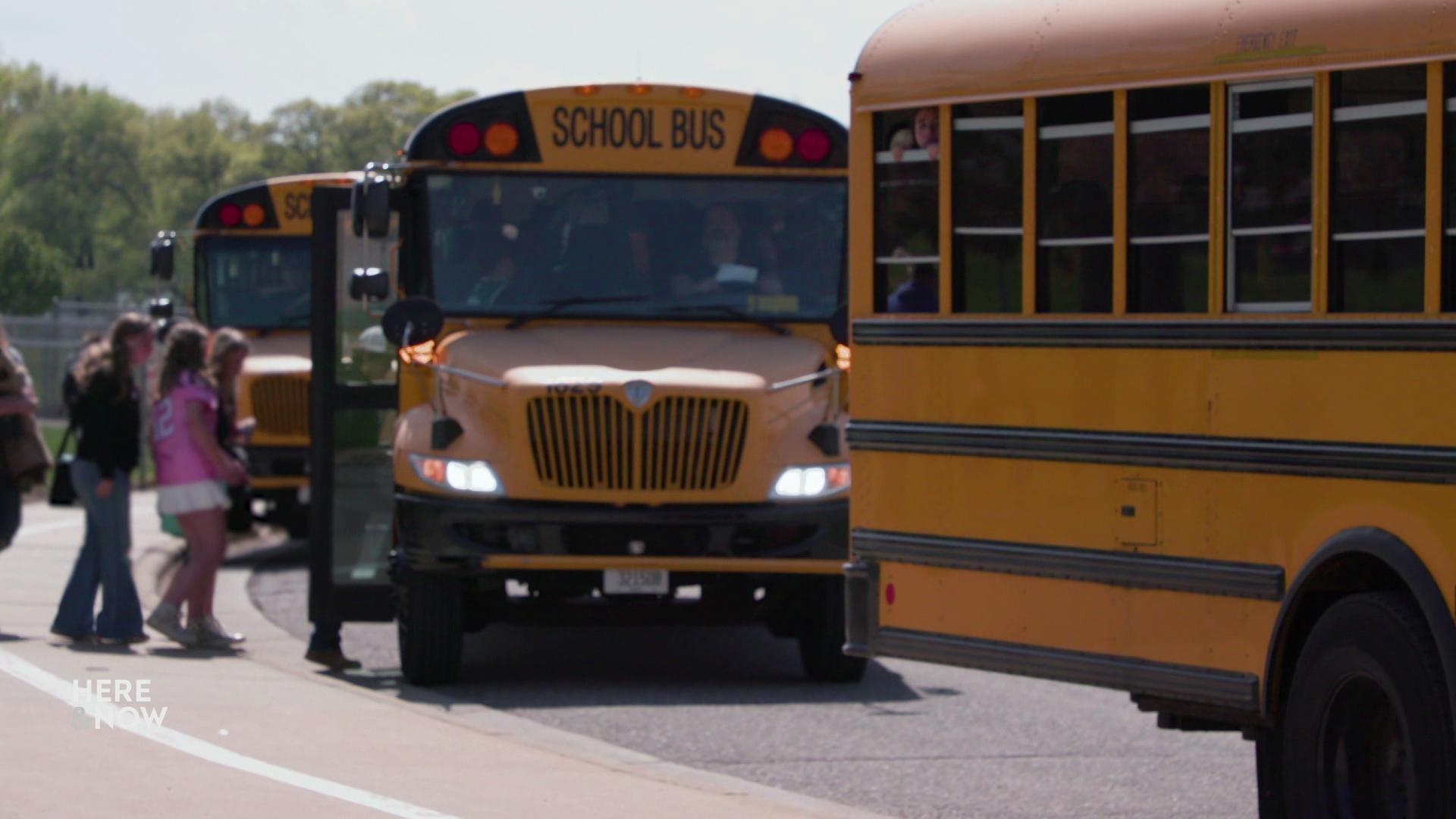 A still image shows a line of school busses picking up students as they depart from school.