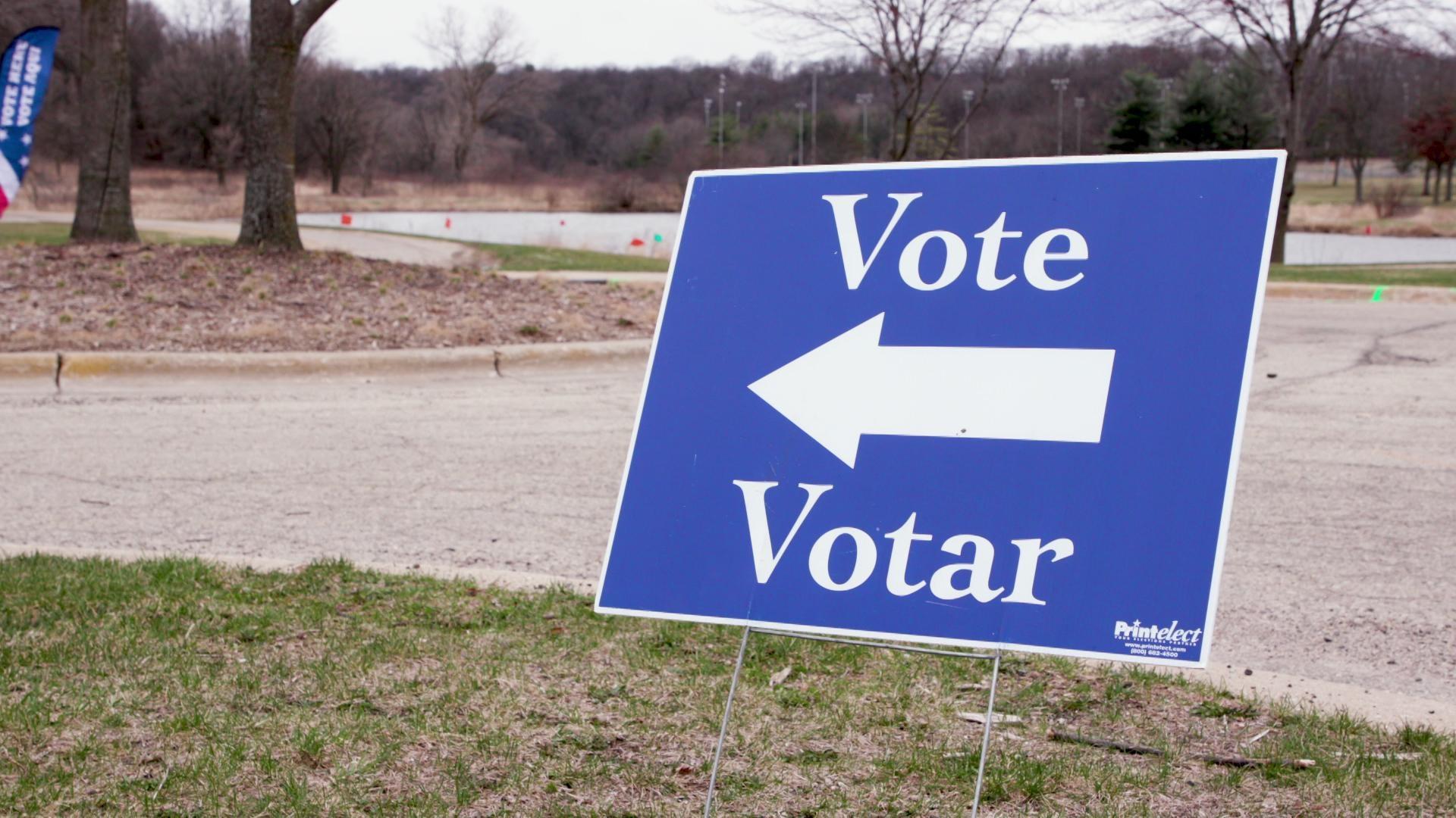 A still image shows a blue yard sign with an arrow pointing to the left, with text surrounding the arrow reading Vote and Votar.