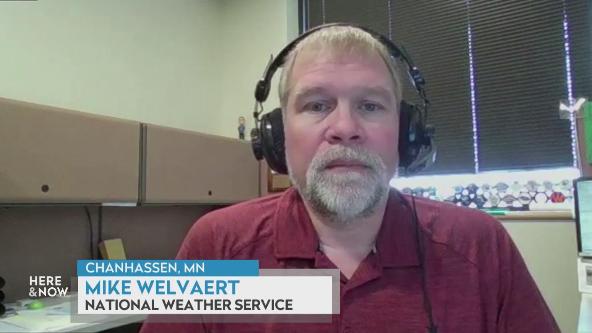 A still image from a video shows Mike Welvaert wearing headphones and sitting in an office in front of cabinets and a window, with a graphic at bottom reading 'Chanhassen, MN,' 'Mike Welvaert' and 'National Weather Service.'