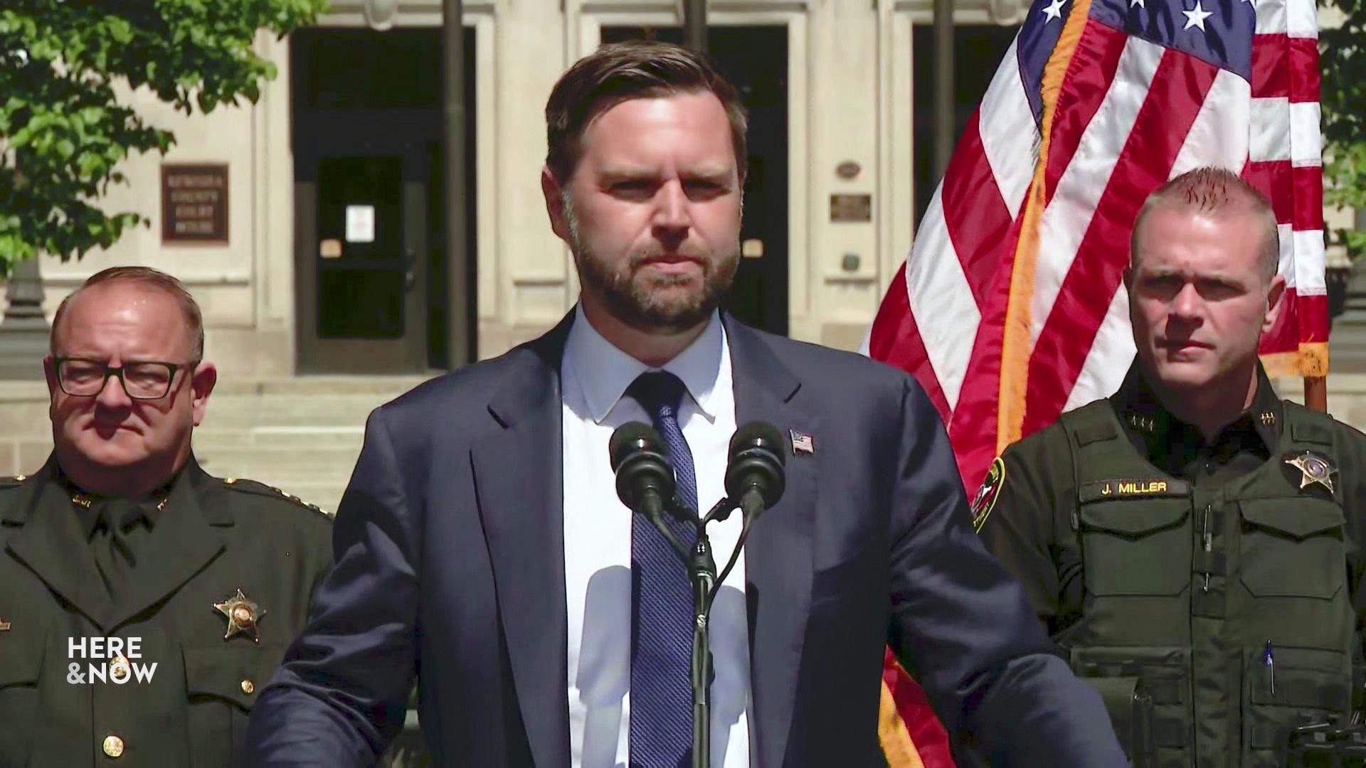 JD Vance stands outside in front of a two microphones with people standing beside him, glass doors and concrete steps and a U.S. flag in the background.