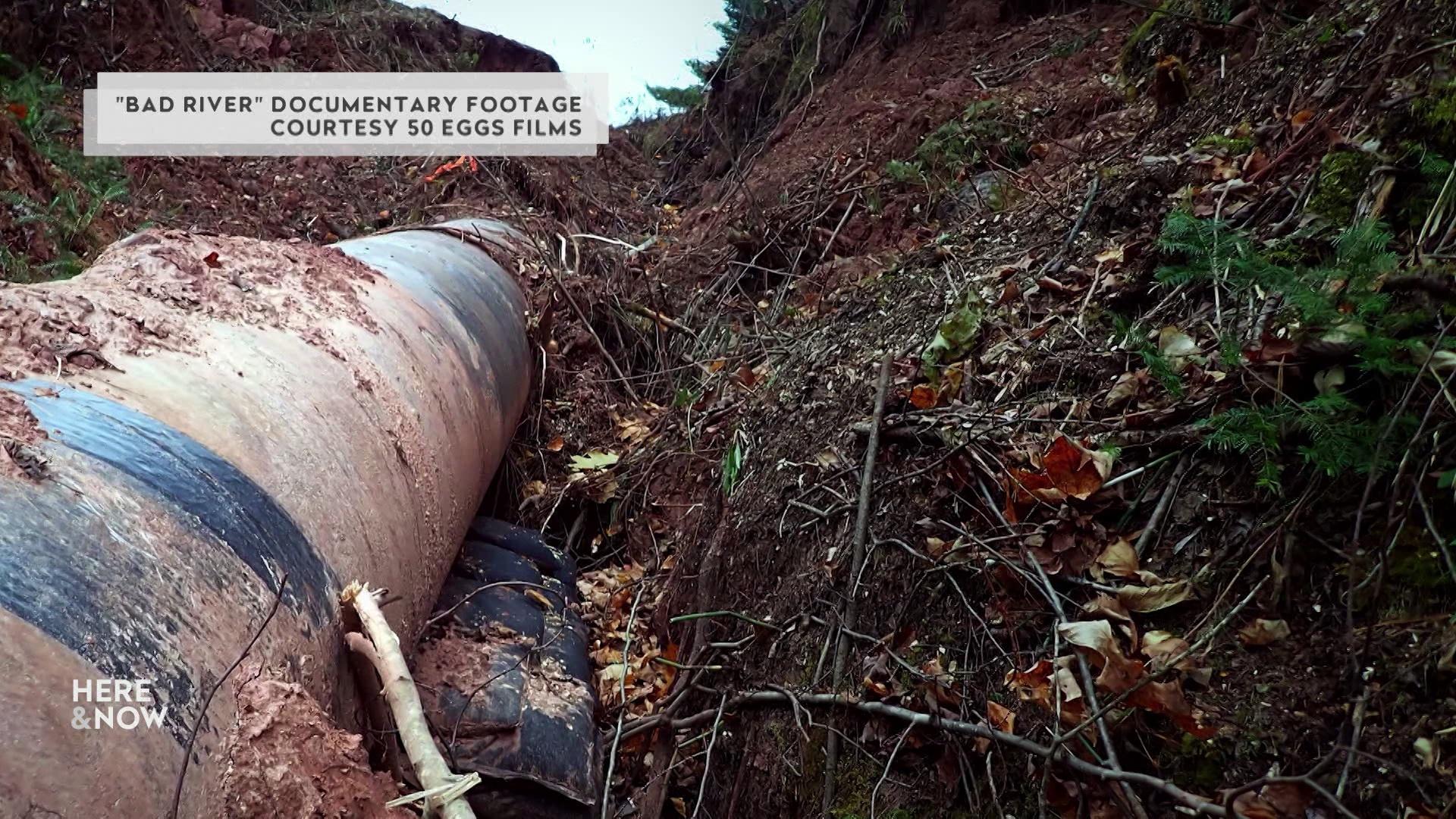 A metal pipe rests on dug up earth with dirt, sticks and debris around it and a graphic at the top reading 'Bad River Documentary Footage Courtesy 50 Eggs Films.'