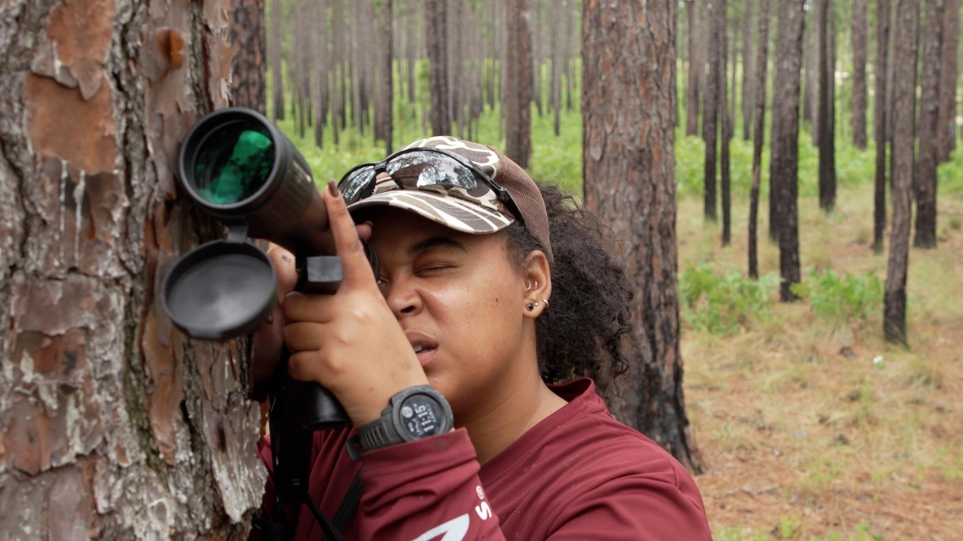 A Black woman in the woods, looking through a telescope, birding.